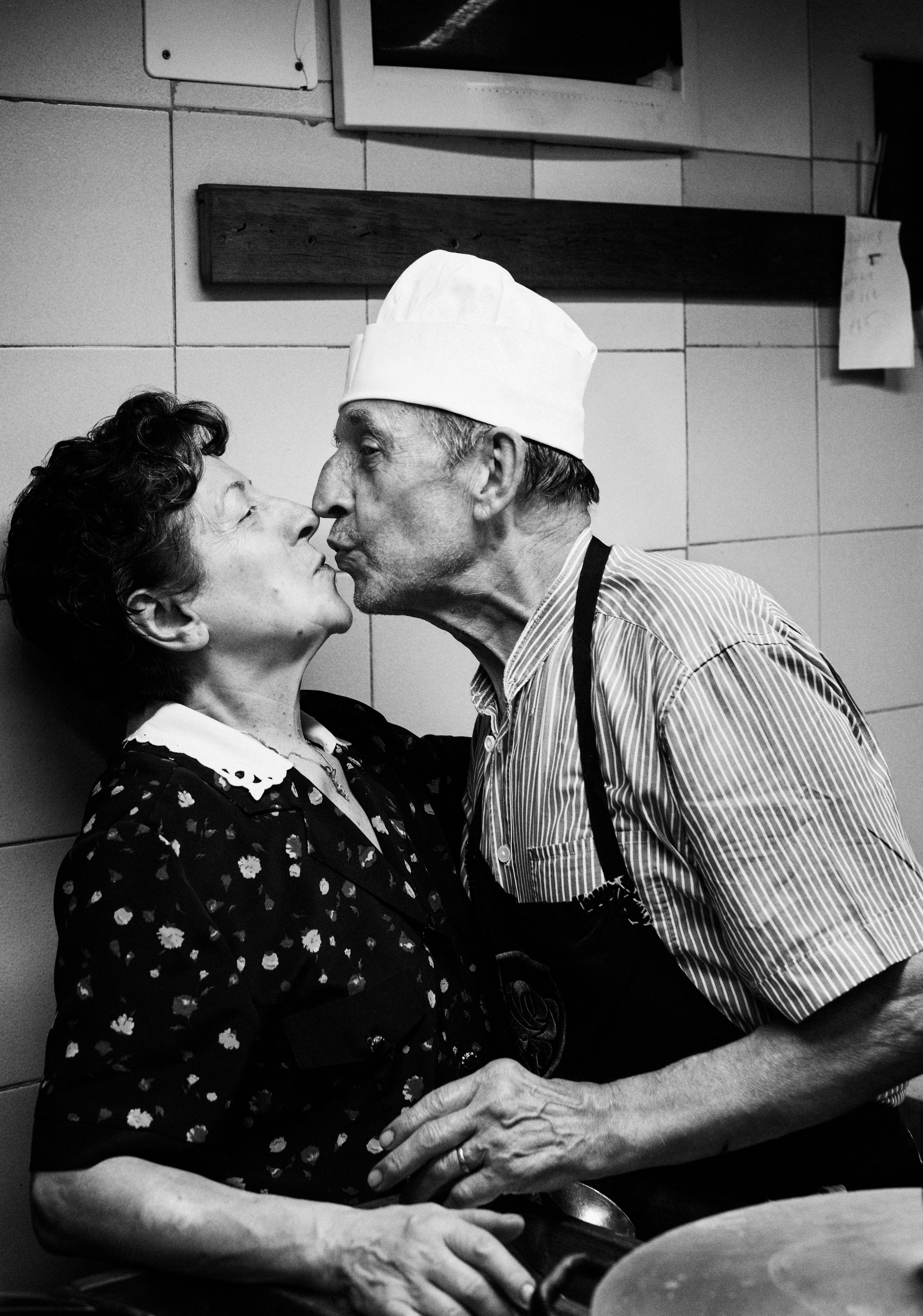 An elderly man wearing a chef’s hat kisses a woman in a floral dress in a kitchen.