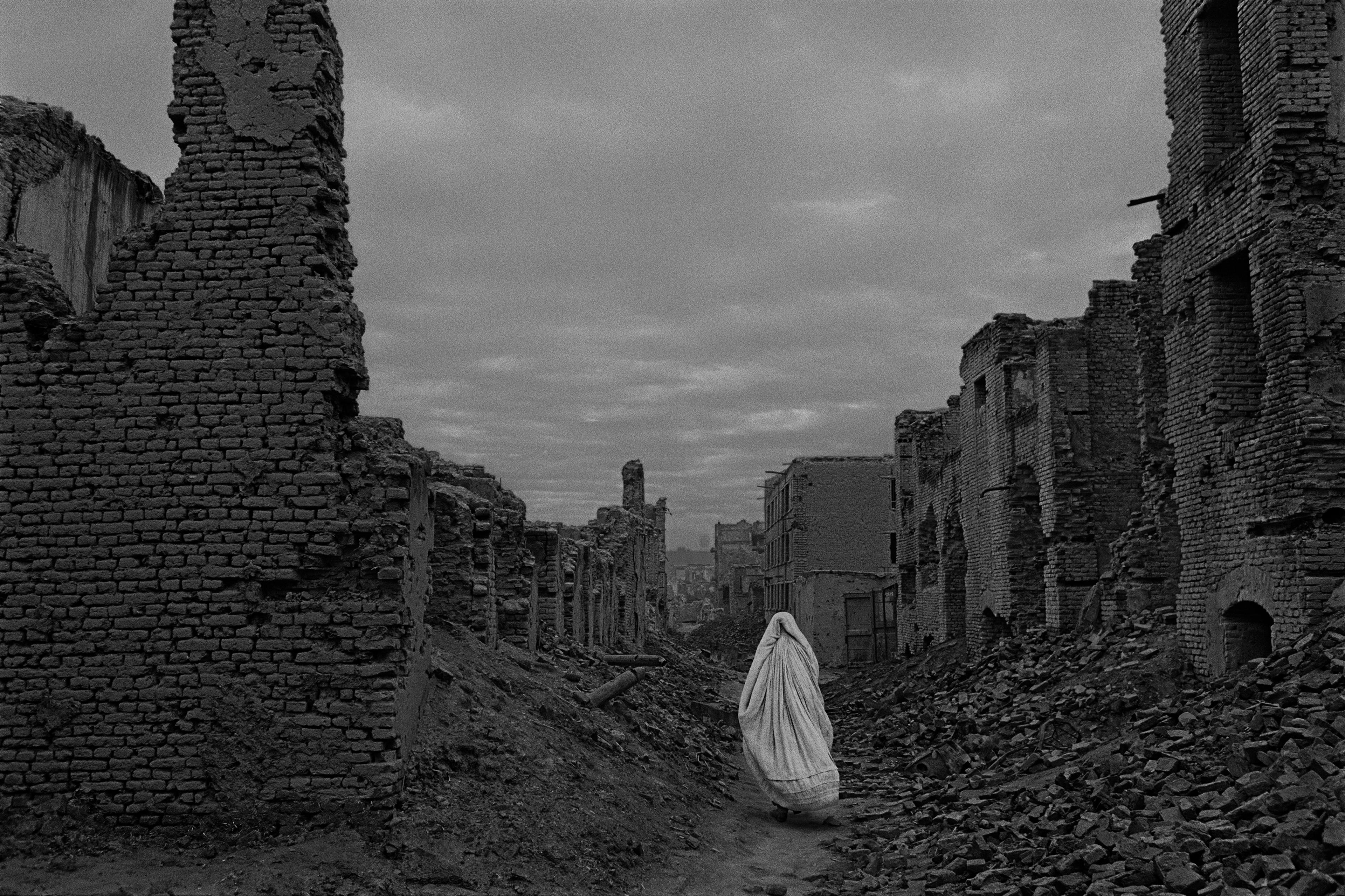 B/w image of a woman with white gown walking away from the viewer in between ruins of houses