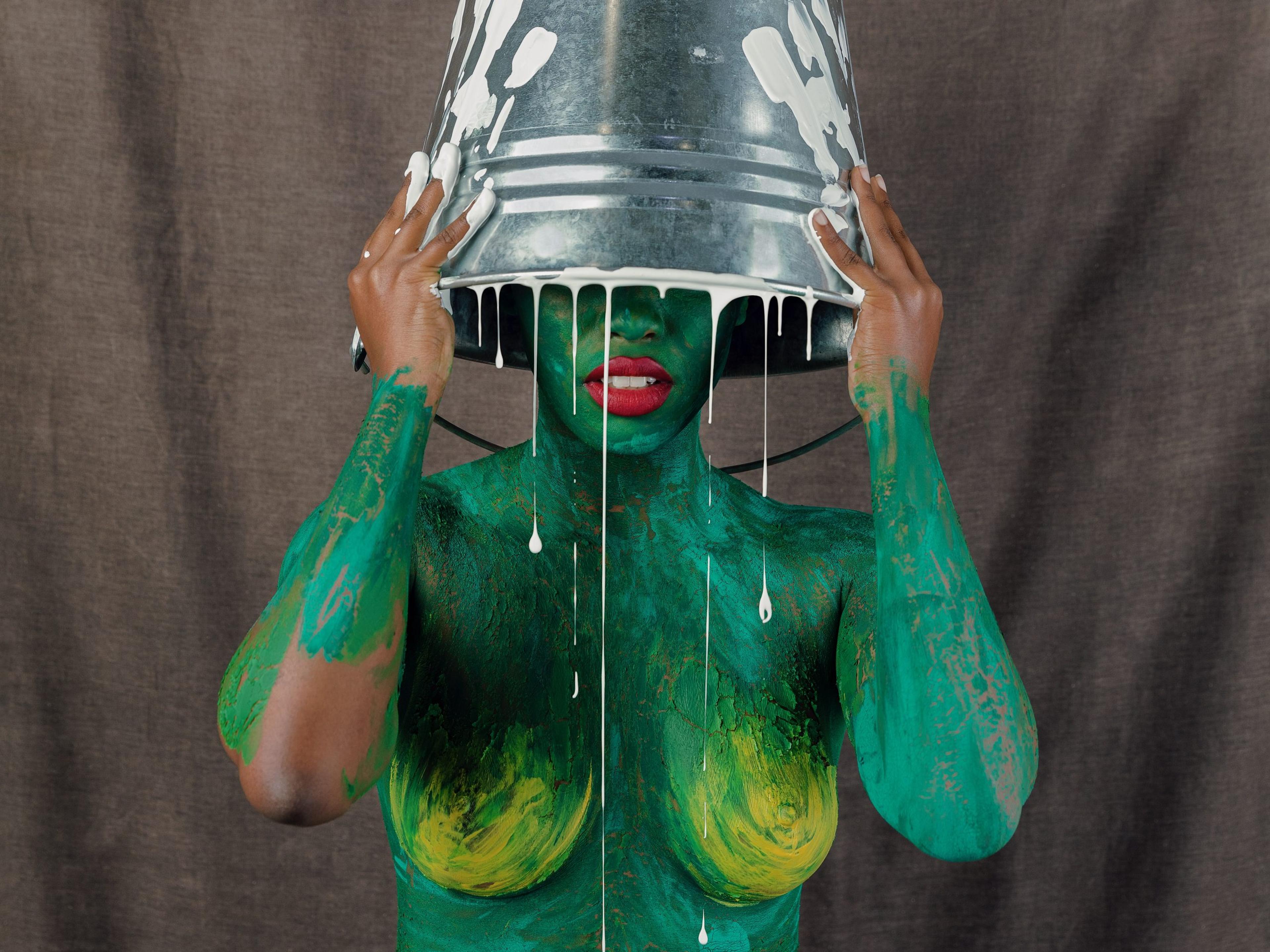 A photograph of a nude woman covered in green paint, holding a dripping paint bucket above her head with both hands