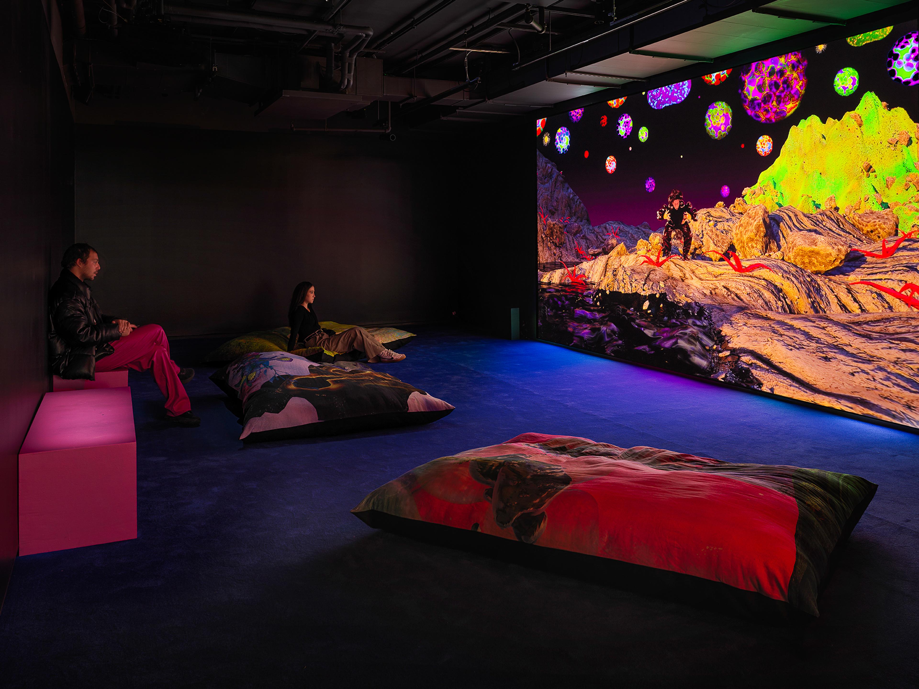 A photograph of a dark room with large cushions and "Matter Gone Wild" by Josèfa Ntjam being projected on a screen