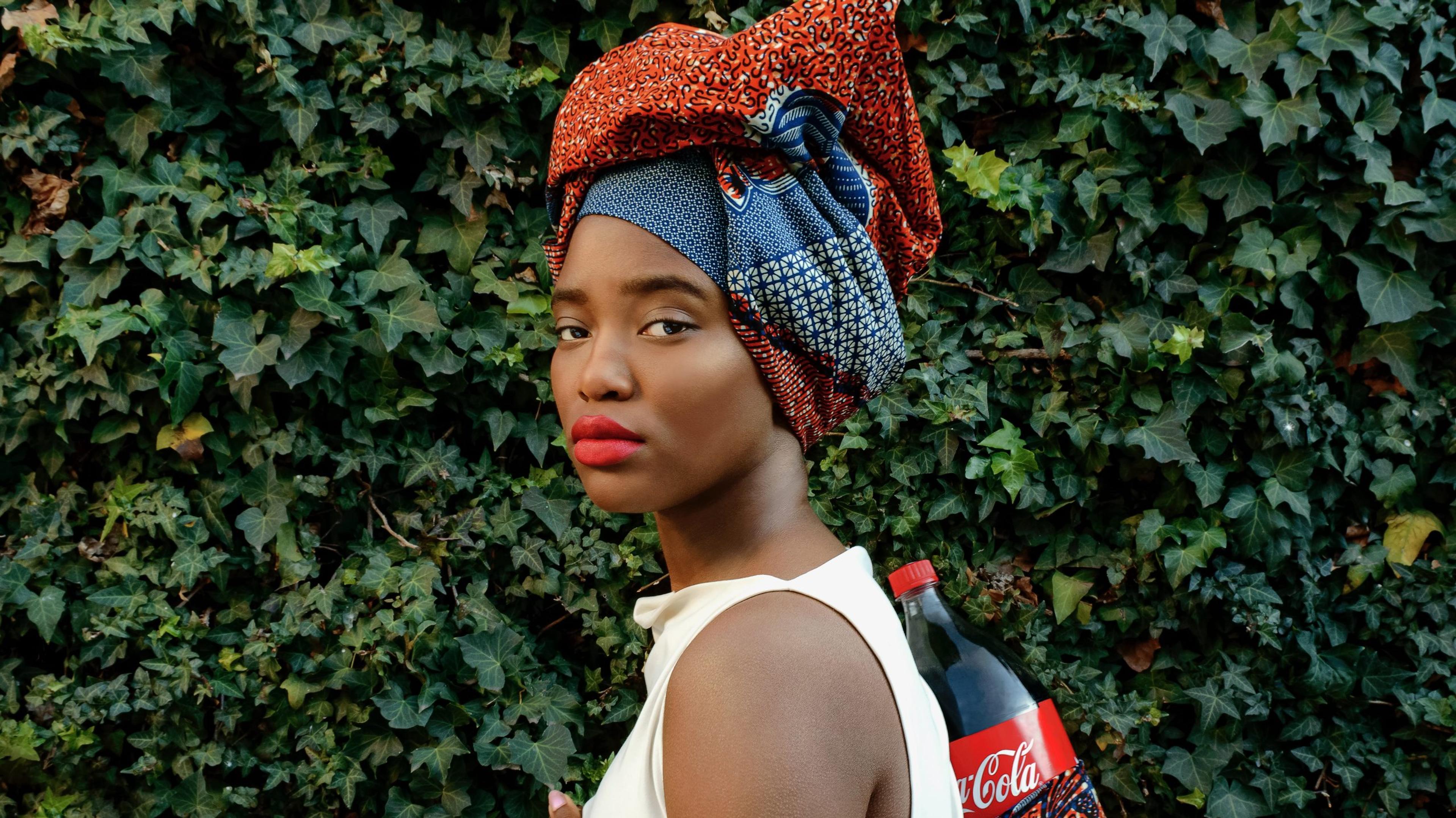 A portrait of a woman wearing red lipstick and a head covering carrying a Coca-Cola bottle on her back, with foliage in the background