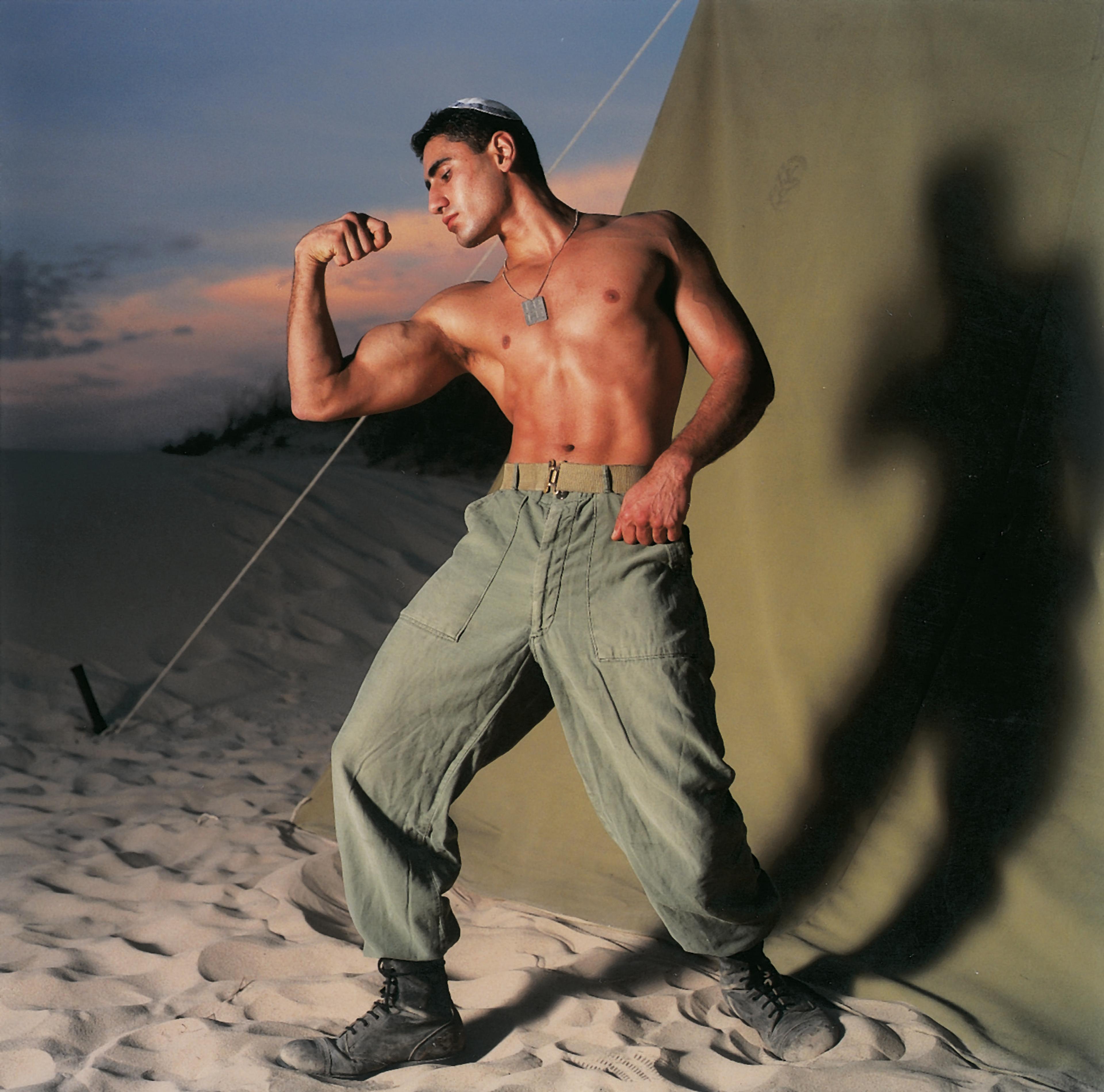 A photograph of a shirtless male soldier in the desert, flexing his bicep