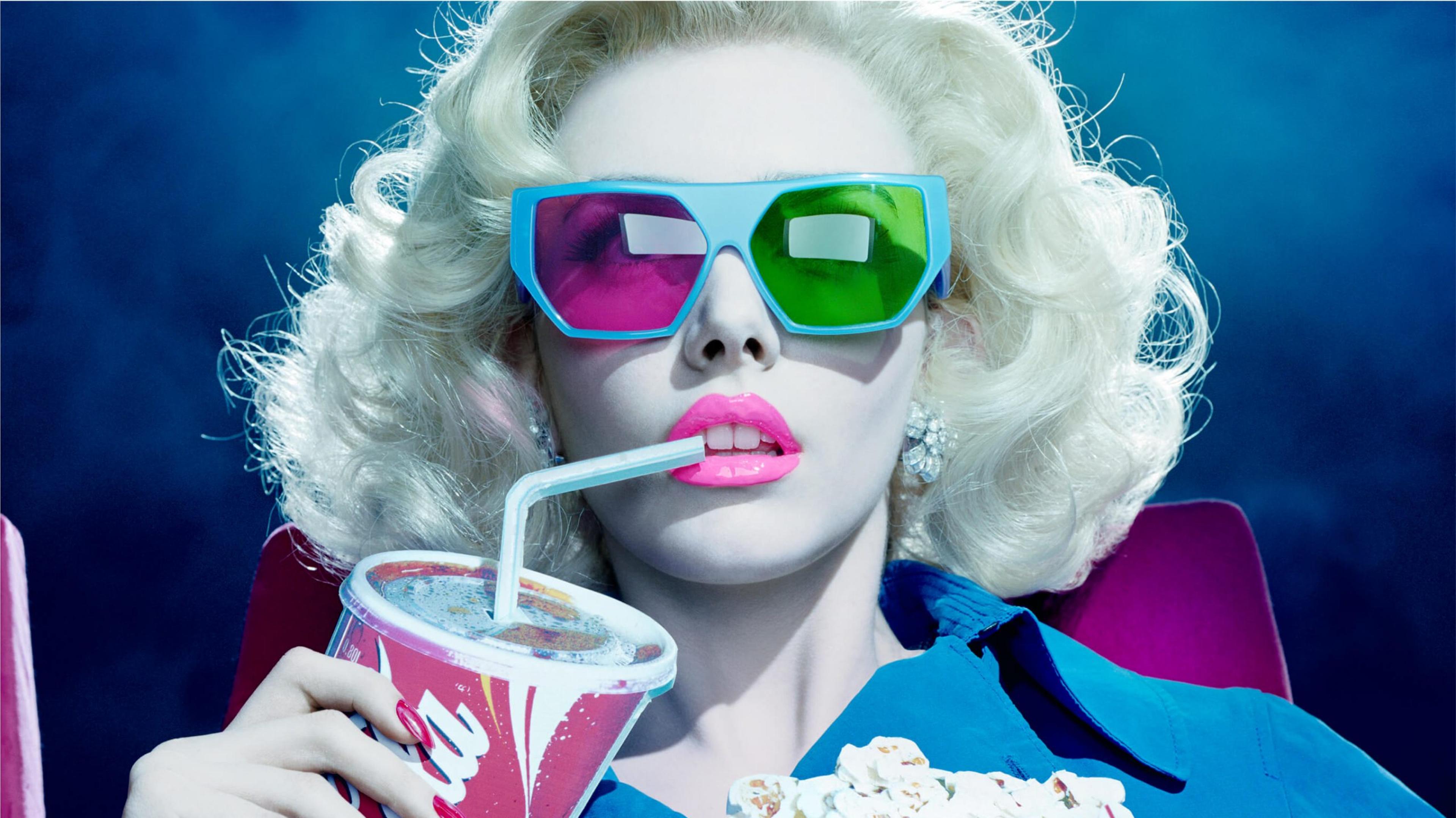 A close-up portrait of a woman with light blonde hair and pink-and-green 3D glasses sitting in a movie theater sipping a Coke