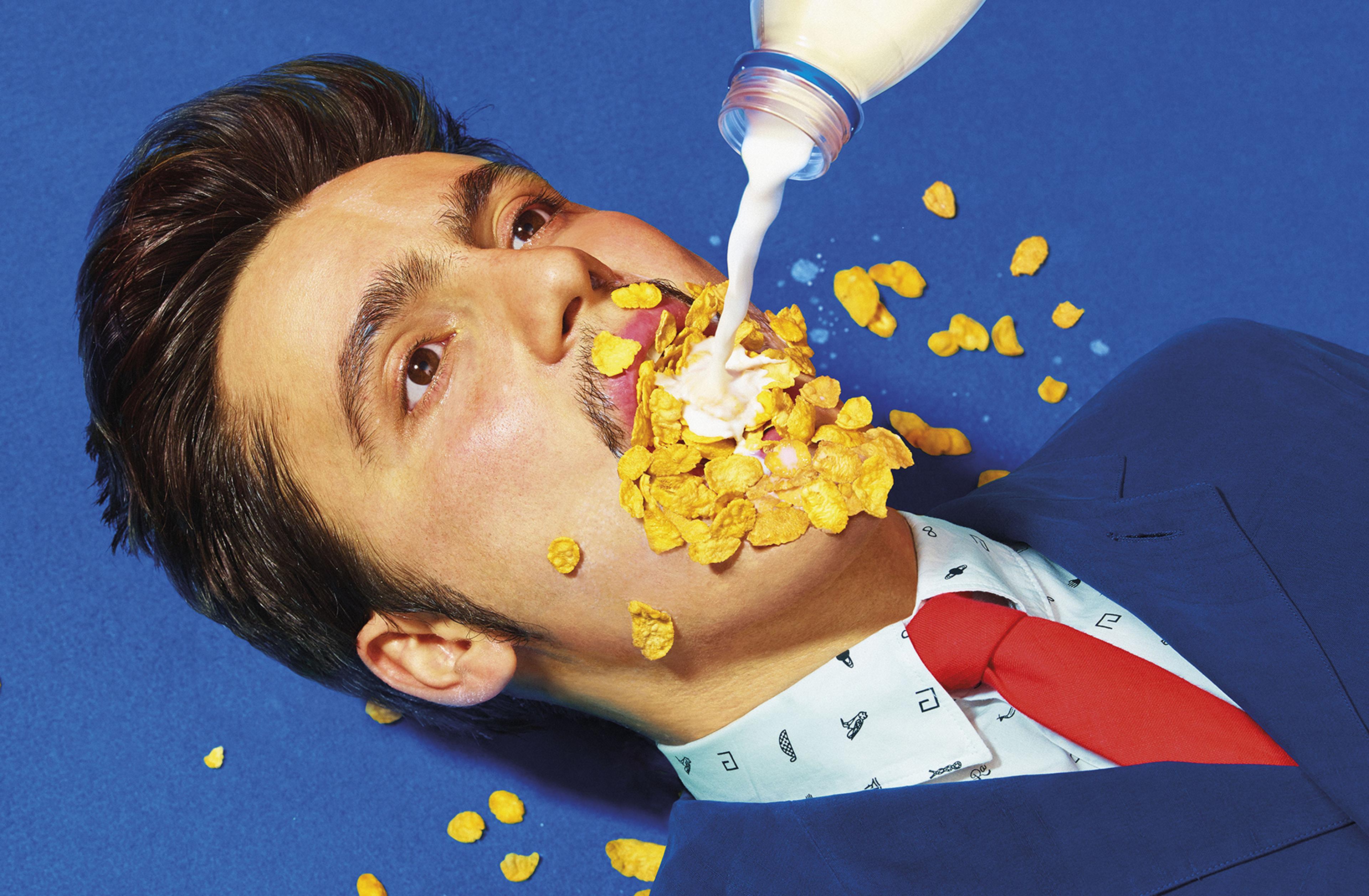 A man in a suit is lying down on a blue floor. he has cornflakes in his mouth and is having milk poured into his mouth