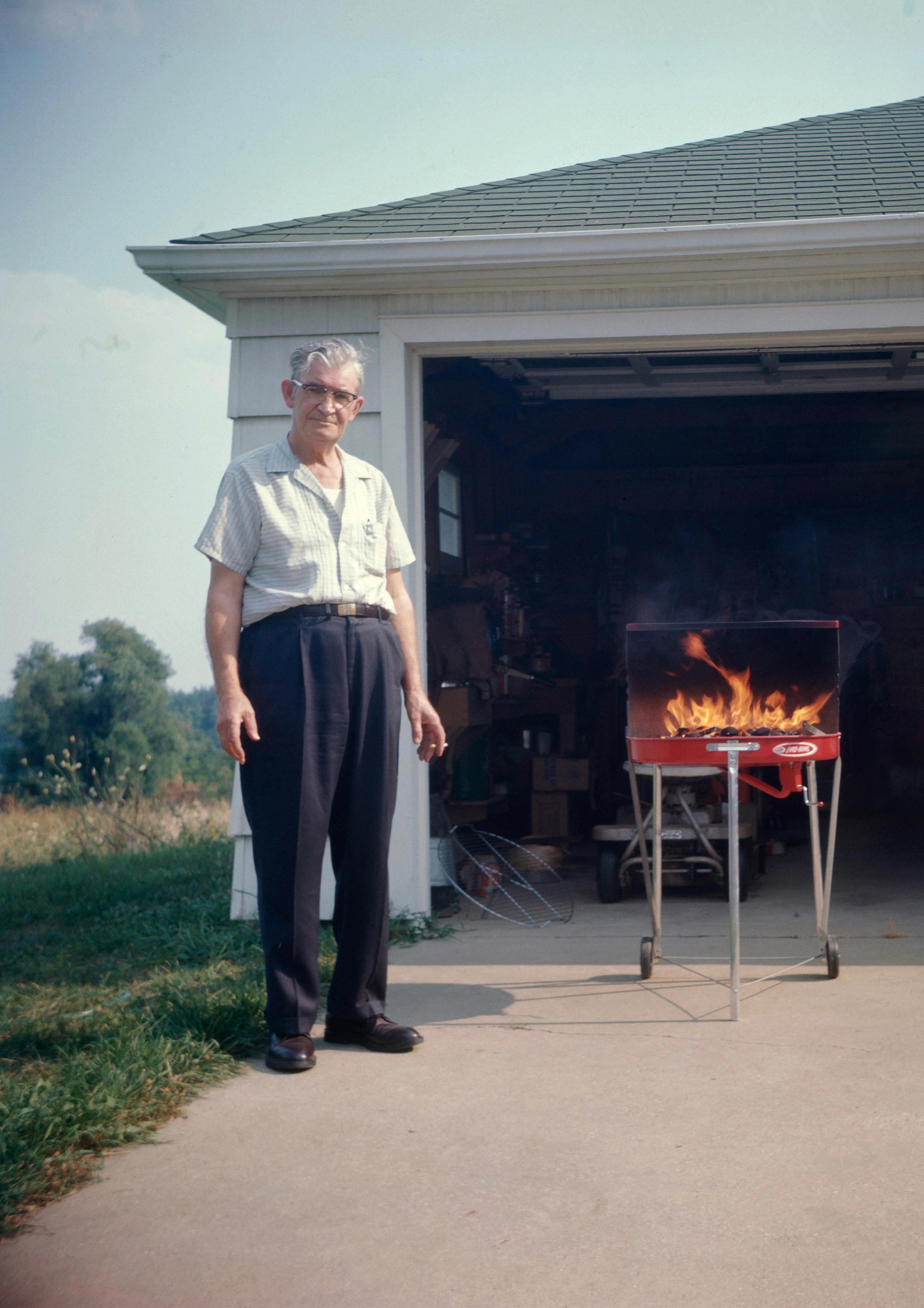 Photograph of a man in the 1950s standing in front of a garage with a grill next to him