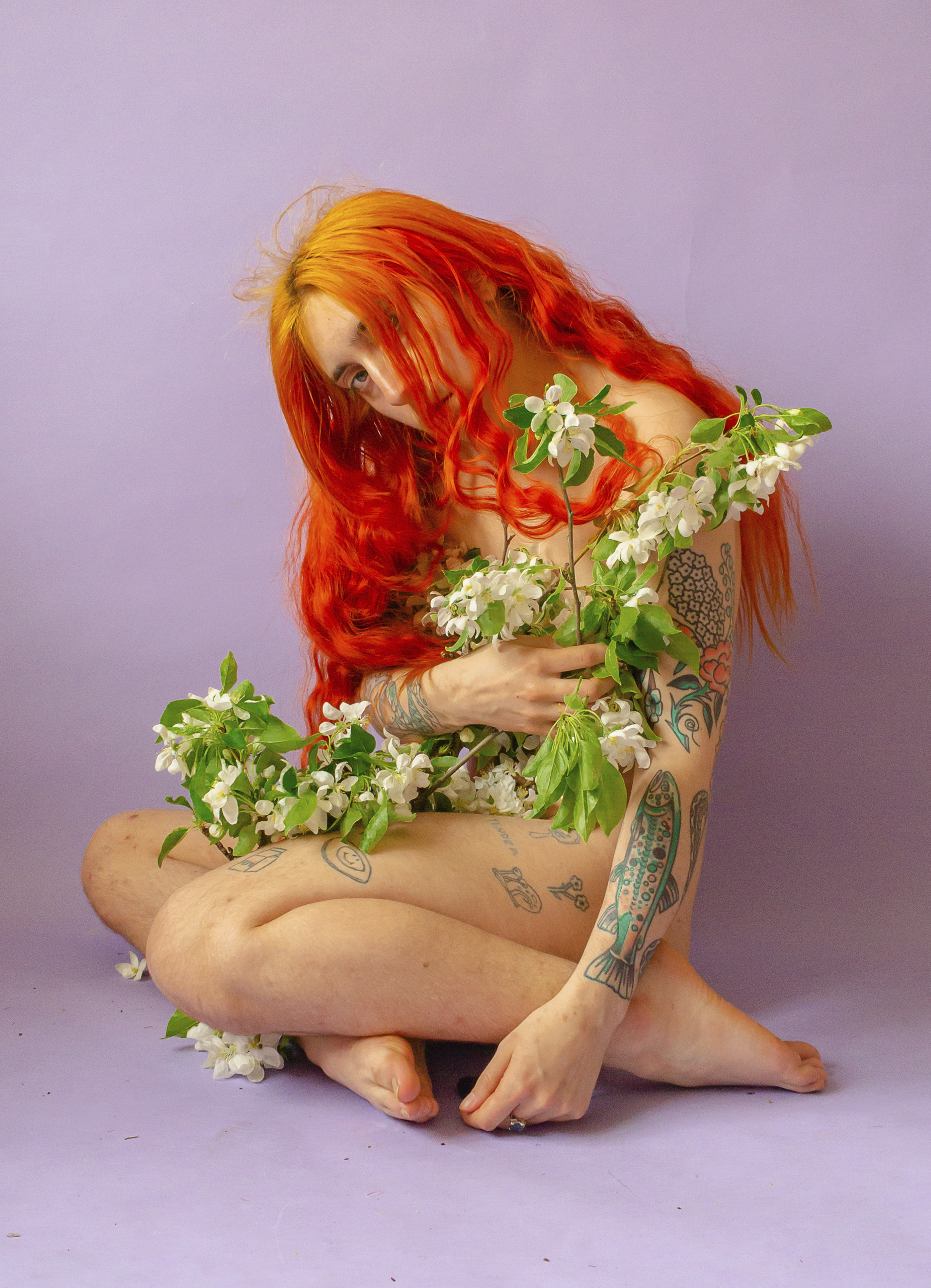 A photograph of a nude red-headed person against a purple background, covering themselves with white flowers