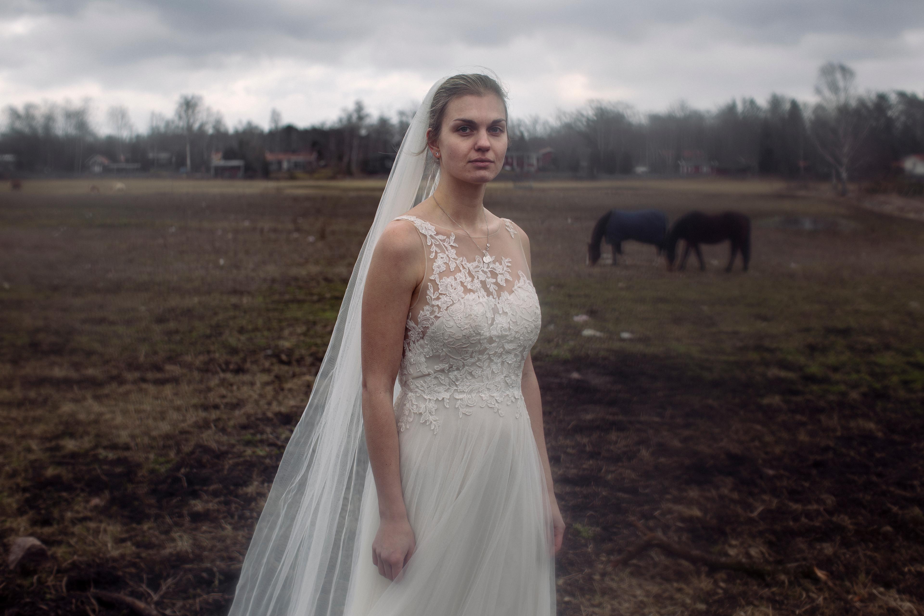 A woman in a wedding dress and veil is standing on a horse field. It is foggy outside and there are two horses in the background