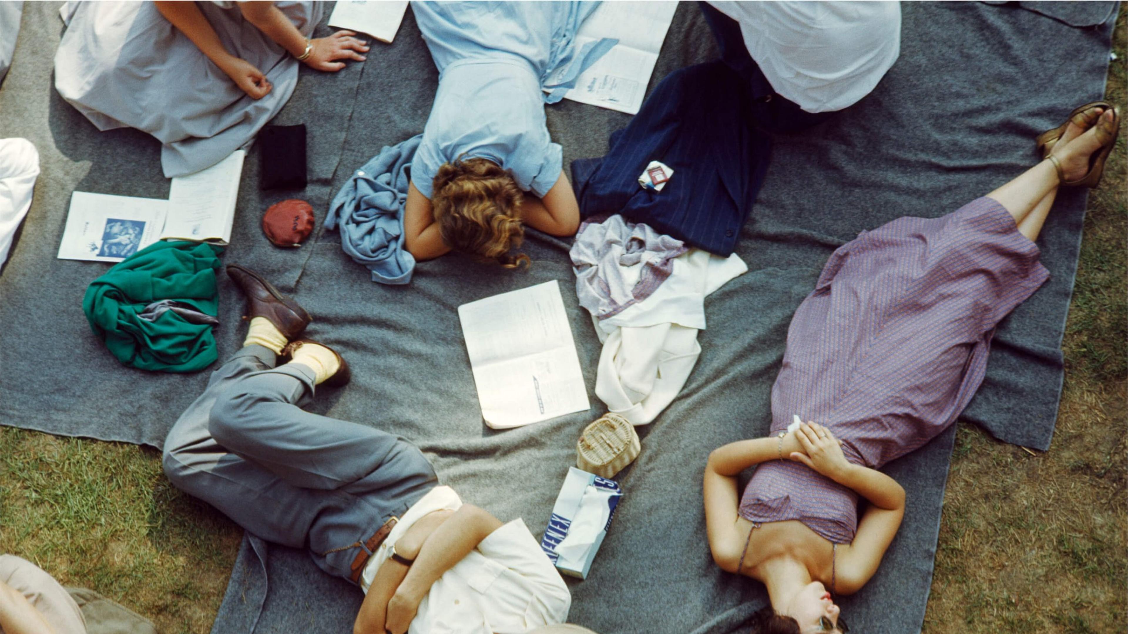 A top-down photograph of a group of people laying down on a grey picnic blanket in the grass