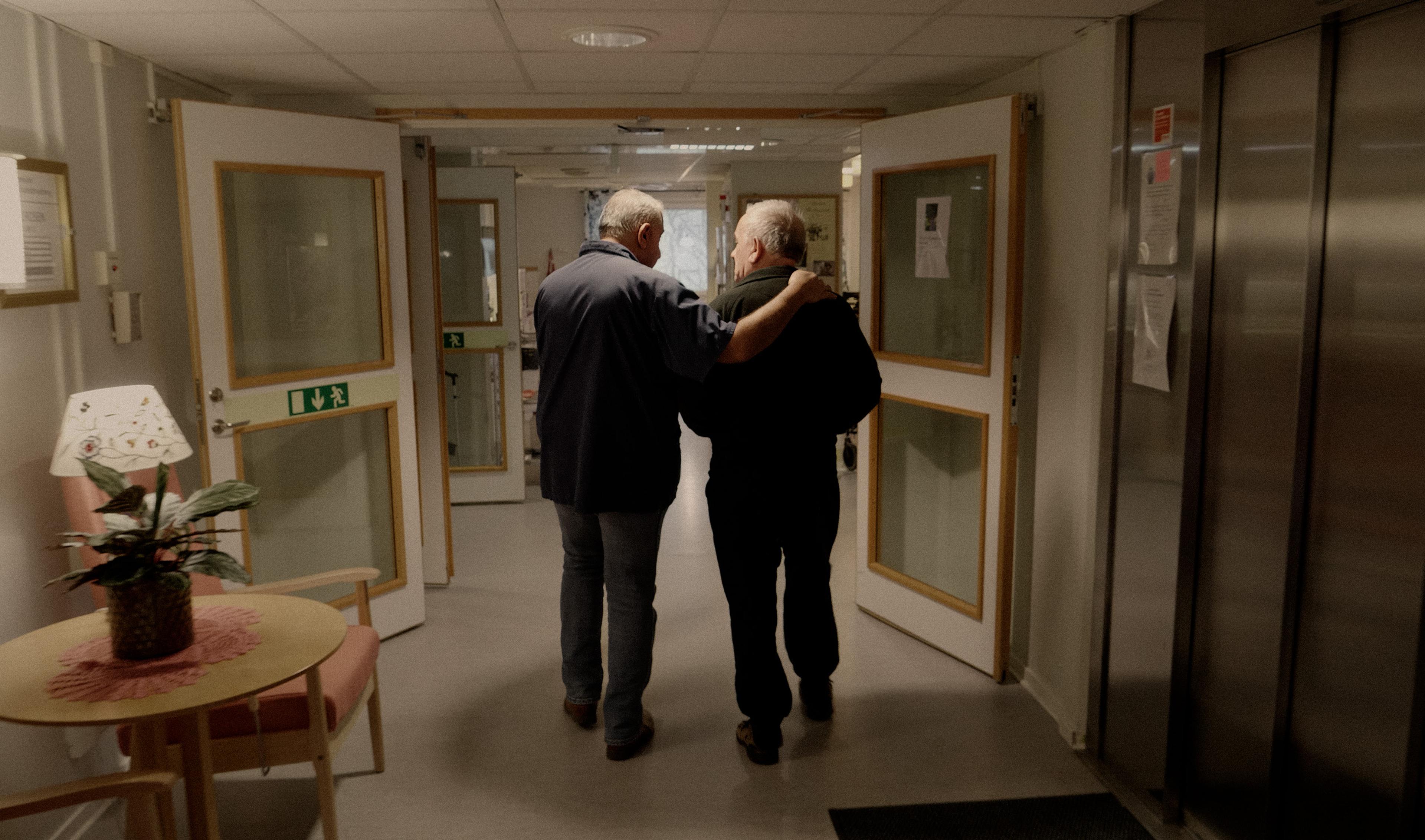 Two men walk together down a hospital or care home corridor, with one putting a supportive arm around the other's shoulders.