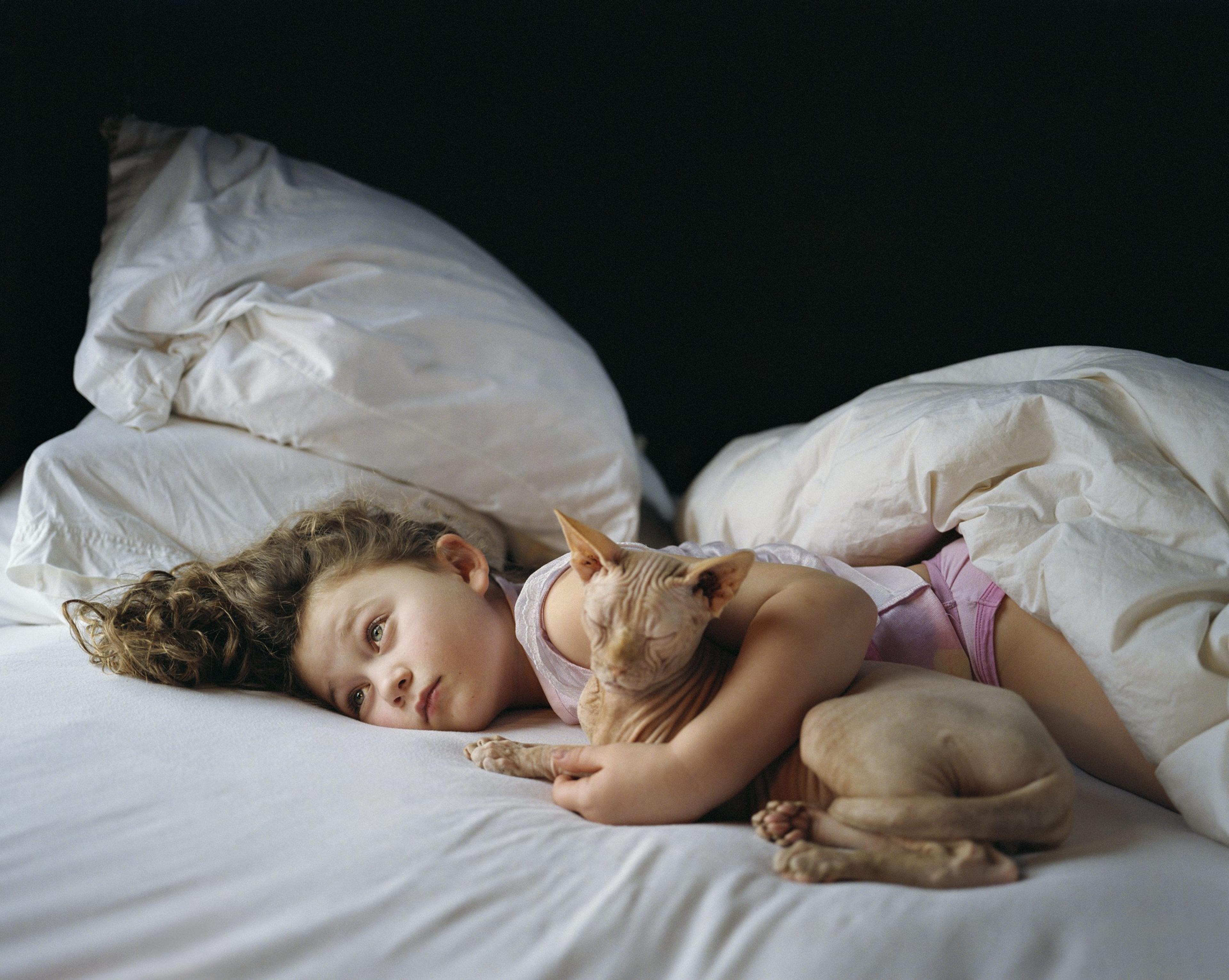 A photograph of a child in bed, hugging a hairless cat