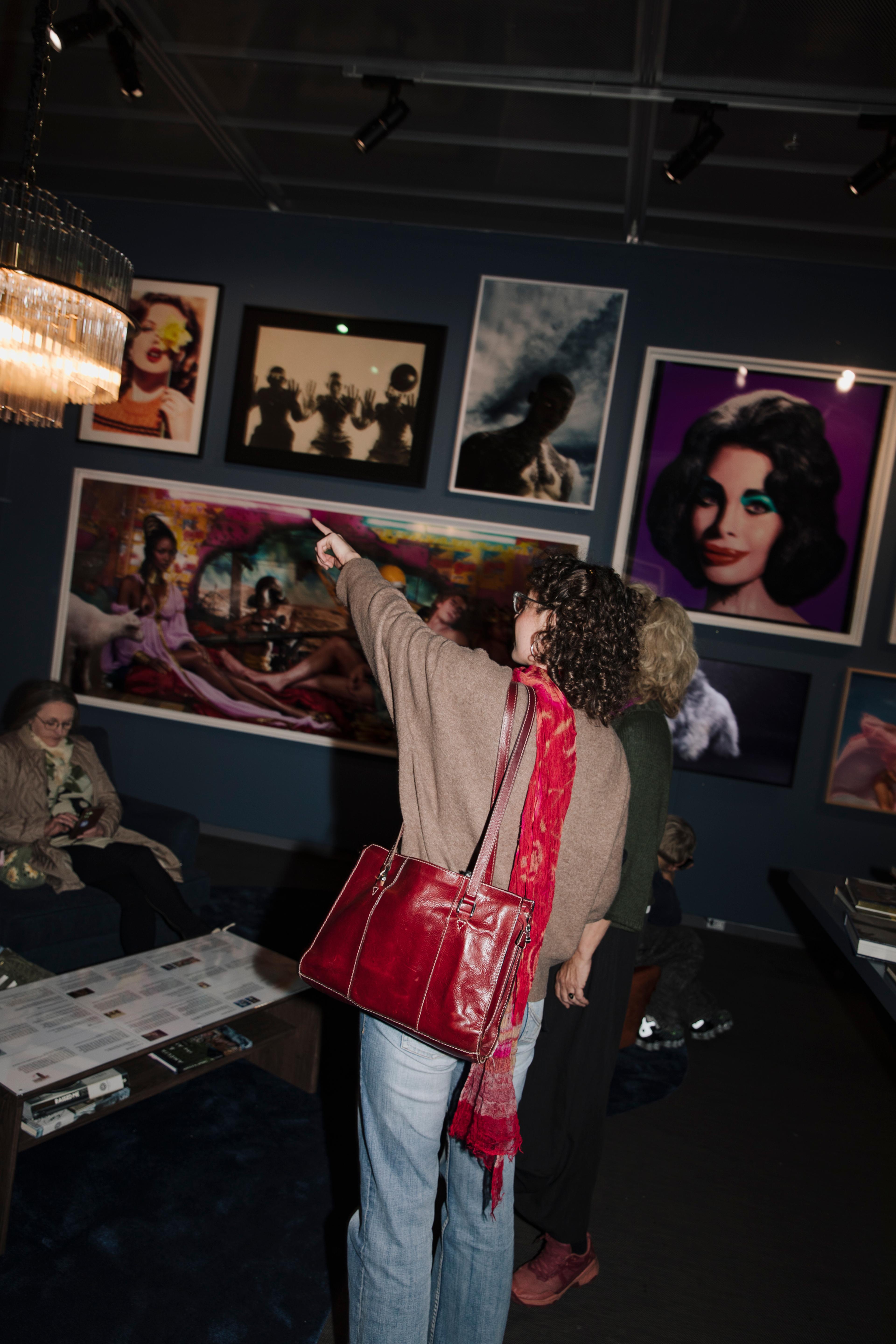 En besökare på ett fotogalleri sedd bakifrån, med en stor röd handväska och röd halsduk. Hon pekar med höger hand mot en av de inramade fotografierna på en mörkblå vägg fylld med olika konstverk.
