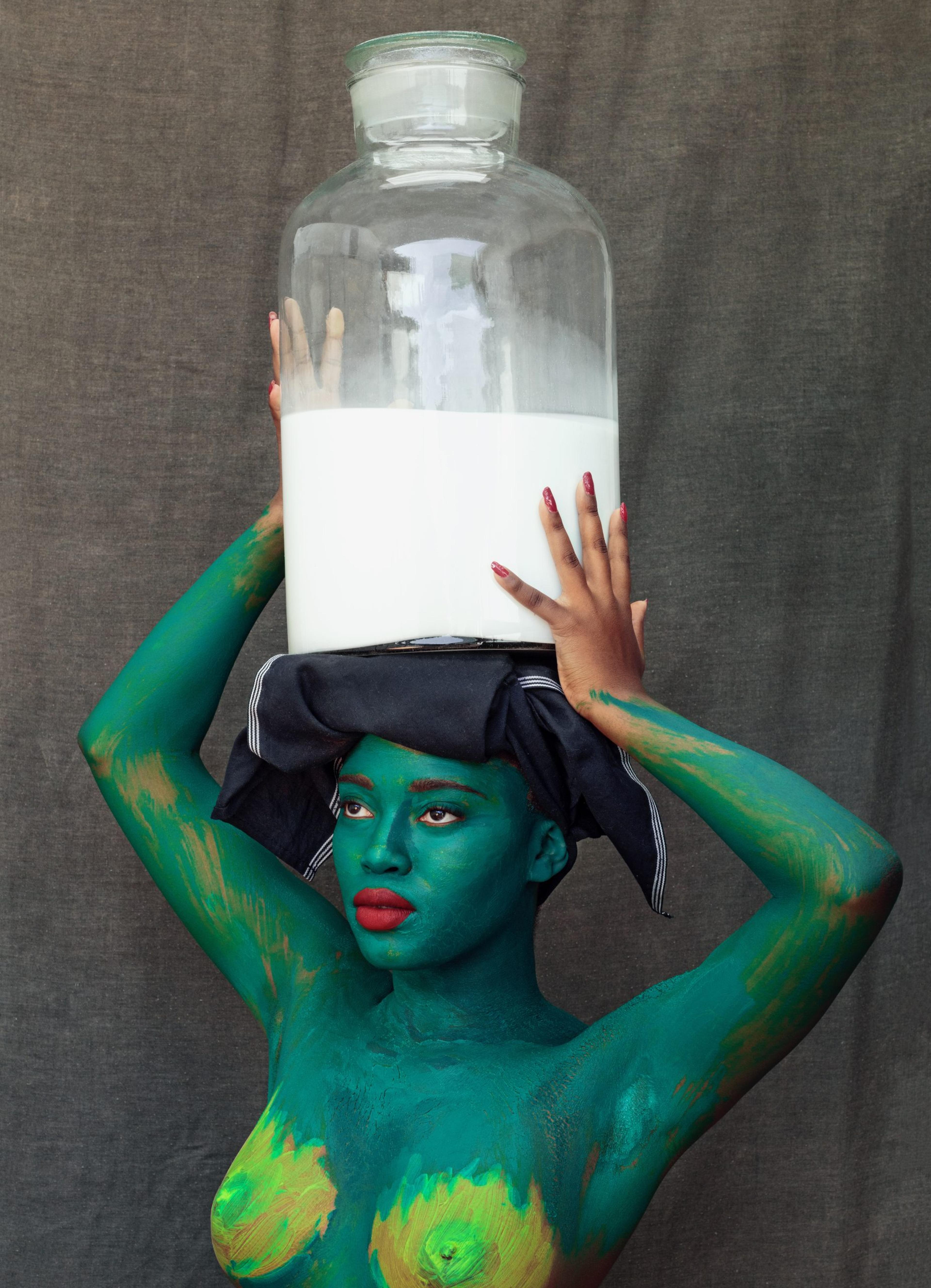 A photograph of a nude woman covered in green paint holding a large vase above her head