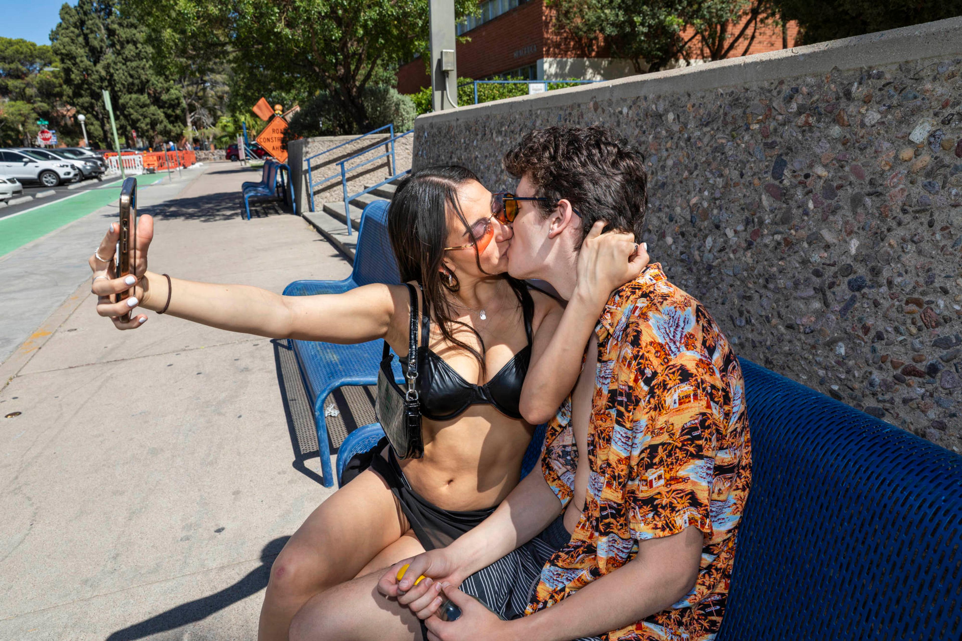 A couple sitting on a bench, kissing, and taking a selfie.