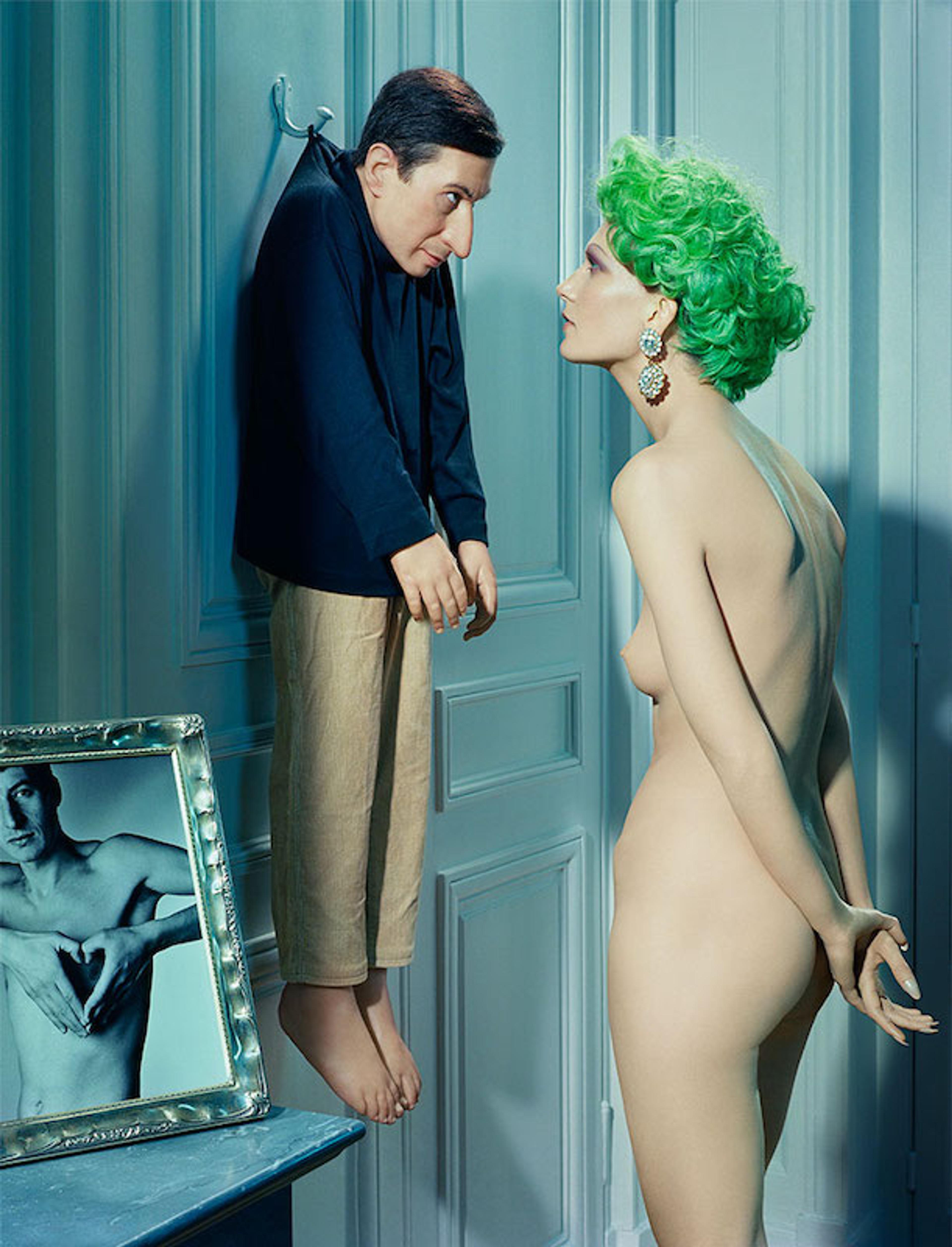 A photograph of a small man hanging on the back of a door by his coat and being stared at by a nude woman with short green hair