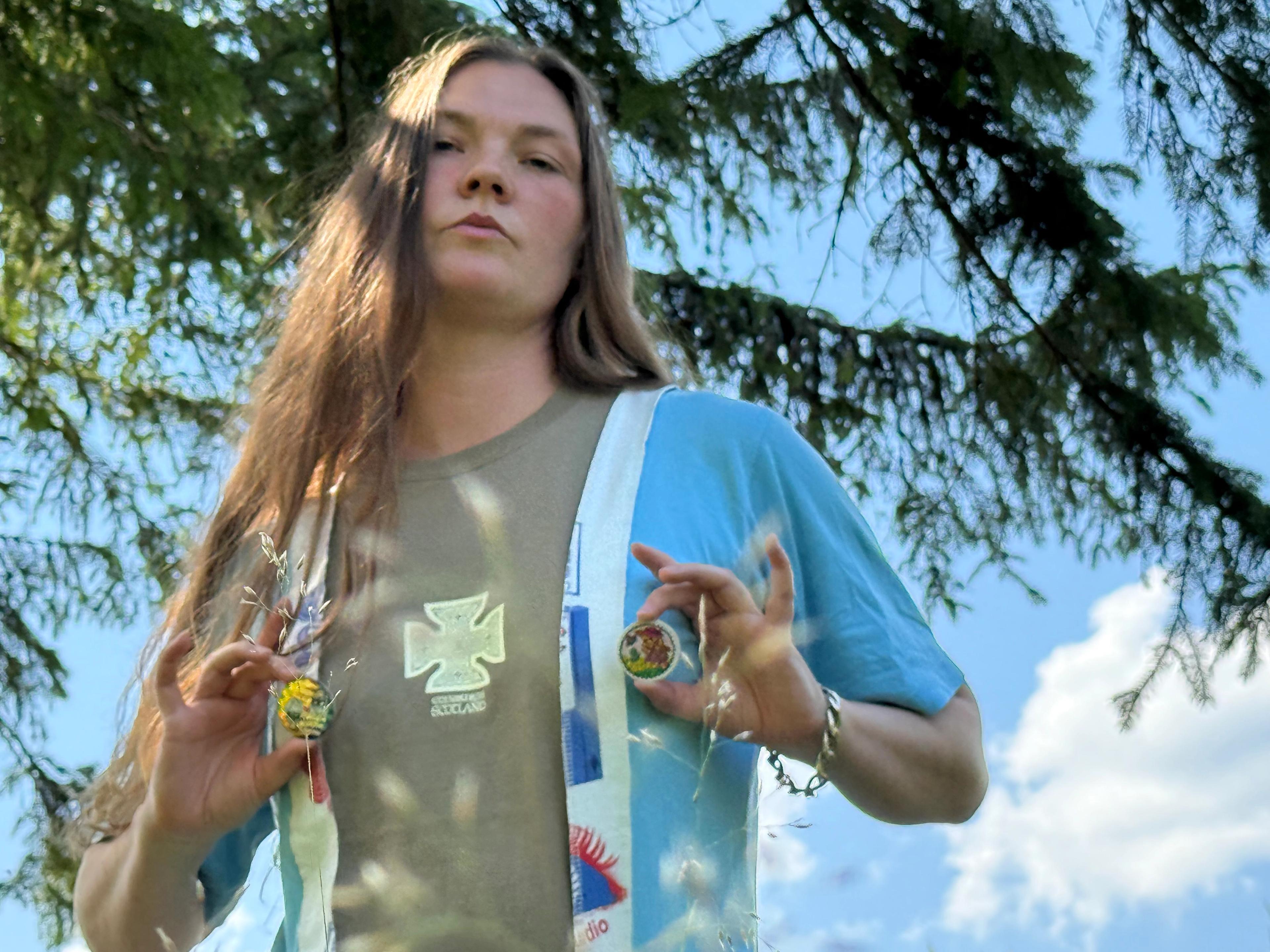 Kvinna med långt hår utomhus som håller upp två små runda föremål framför bröstet, med en granskog och blå himmel i bakgrunden.
