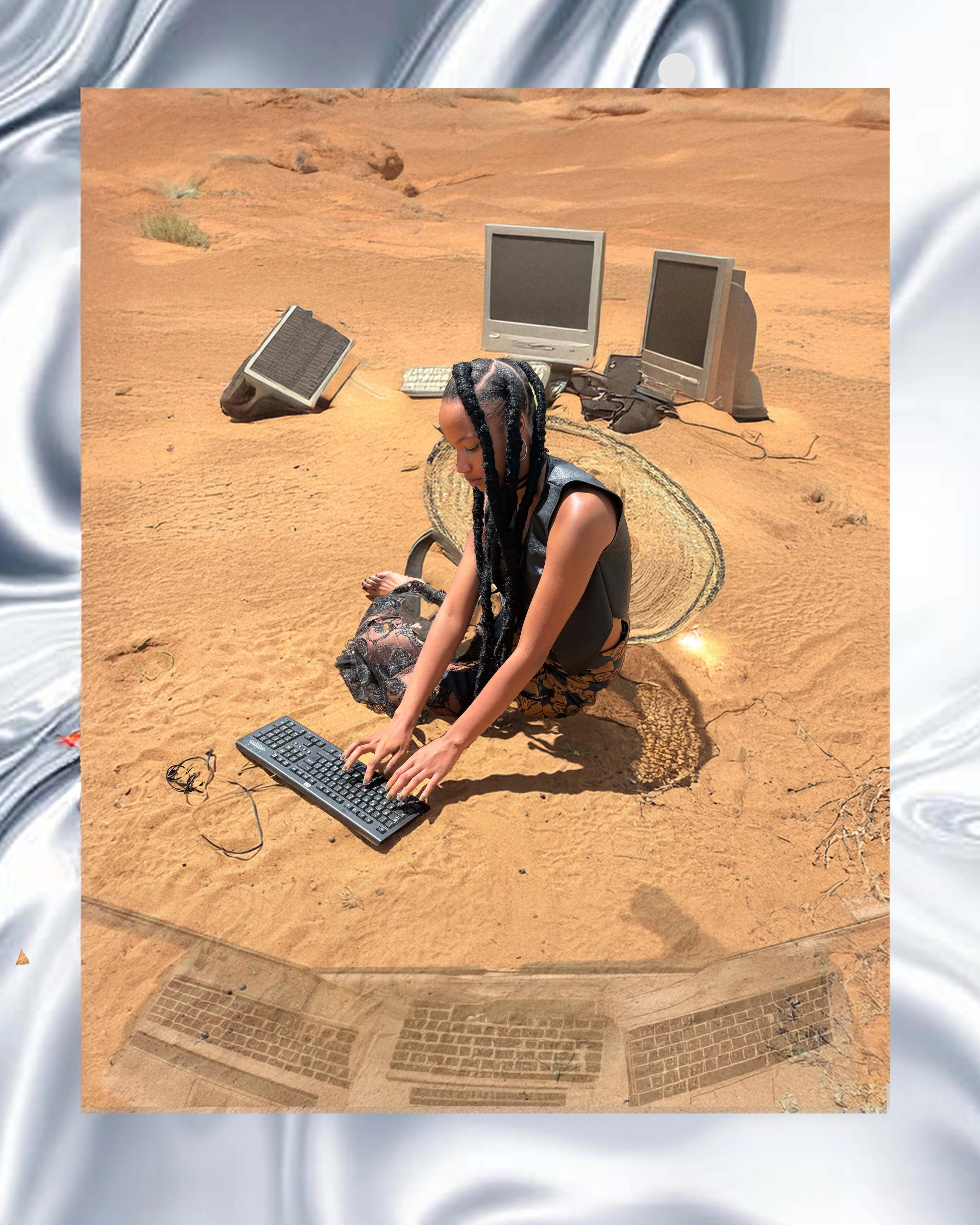 Image of a woman sitting on a sand floor typing on a computer keyboard
