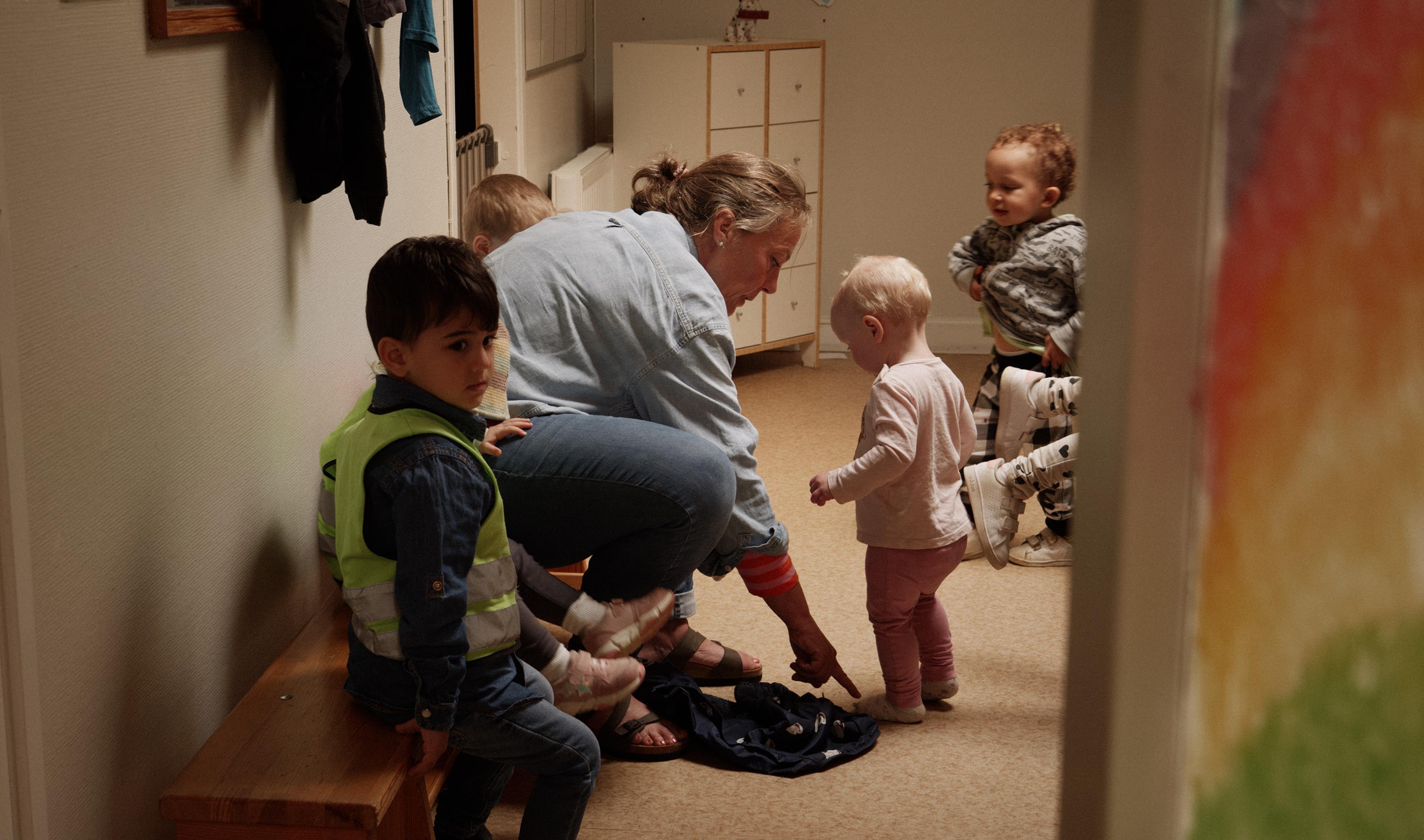 A photograph taken from a hallway looking into a preschool cloakroom. An adult is kneeling down helping four young children put on outdoor clothes. One child is sitting on a wooden bench while others stand and move around. A brightly-lit, domestic-looking scene.