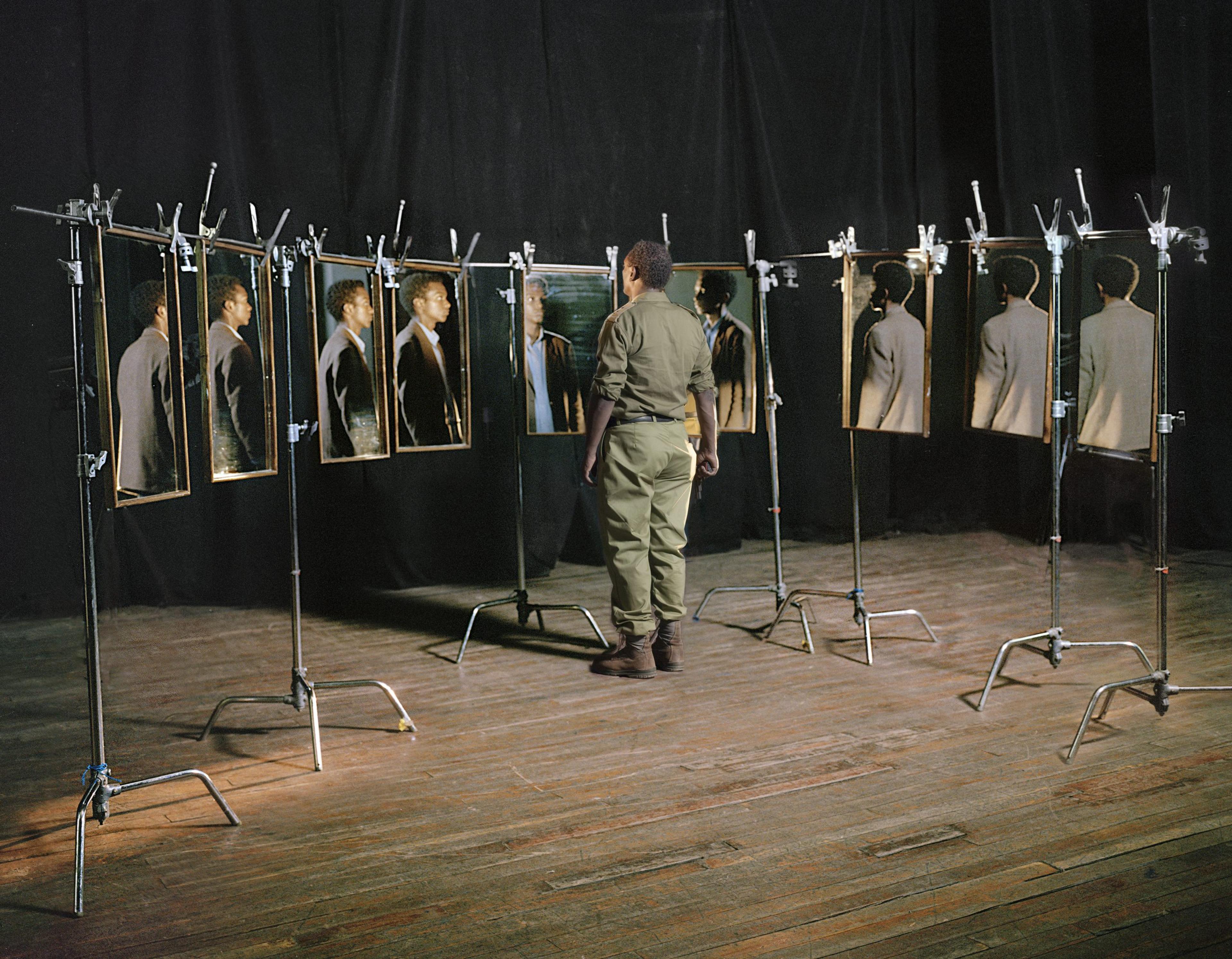 A photograph of a man in military uniform is surrounded by mirrors that reflect back a different man in a brown suit
