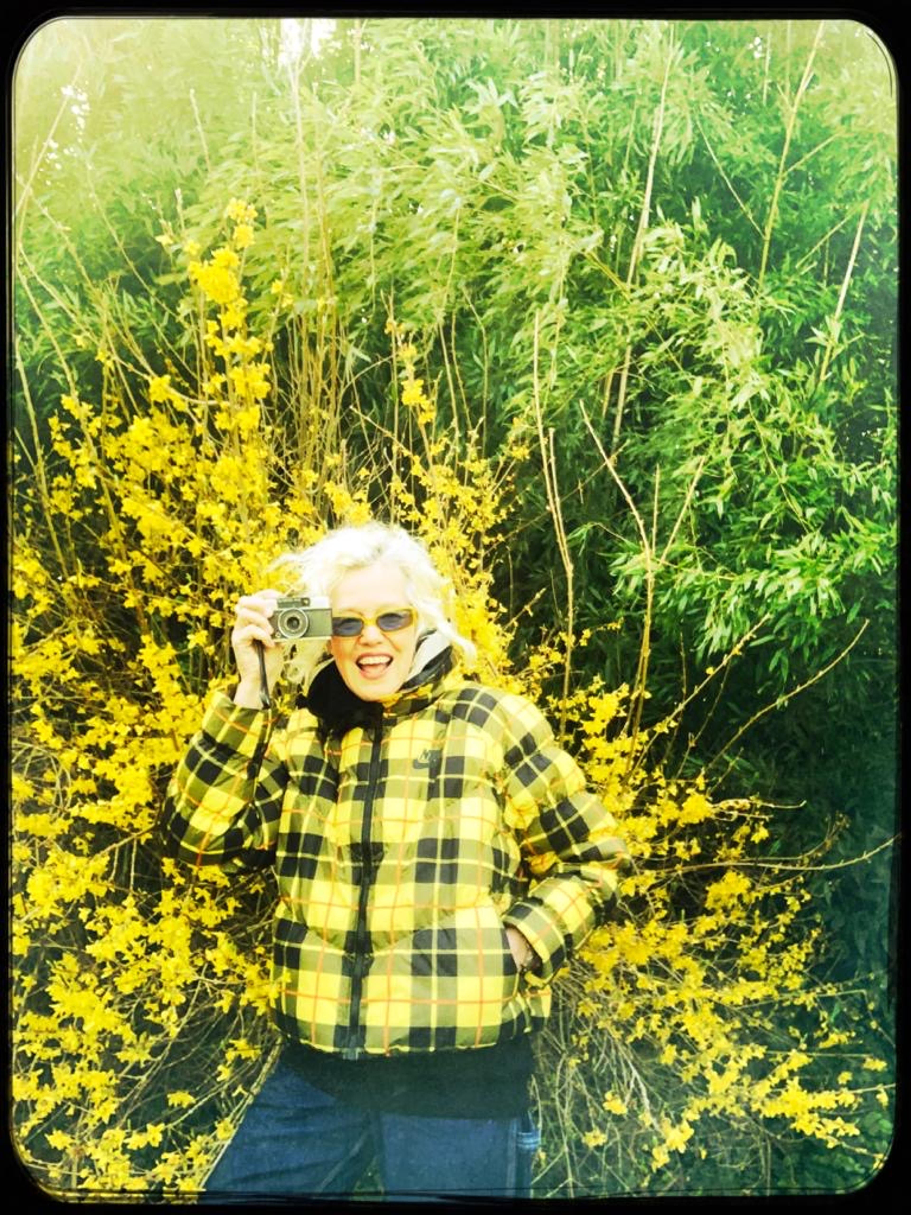 A photograph of a woman in a yellow-and-black plaid jacket holding a camera against a yellow floral background