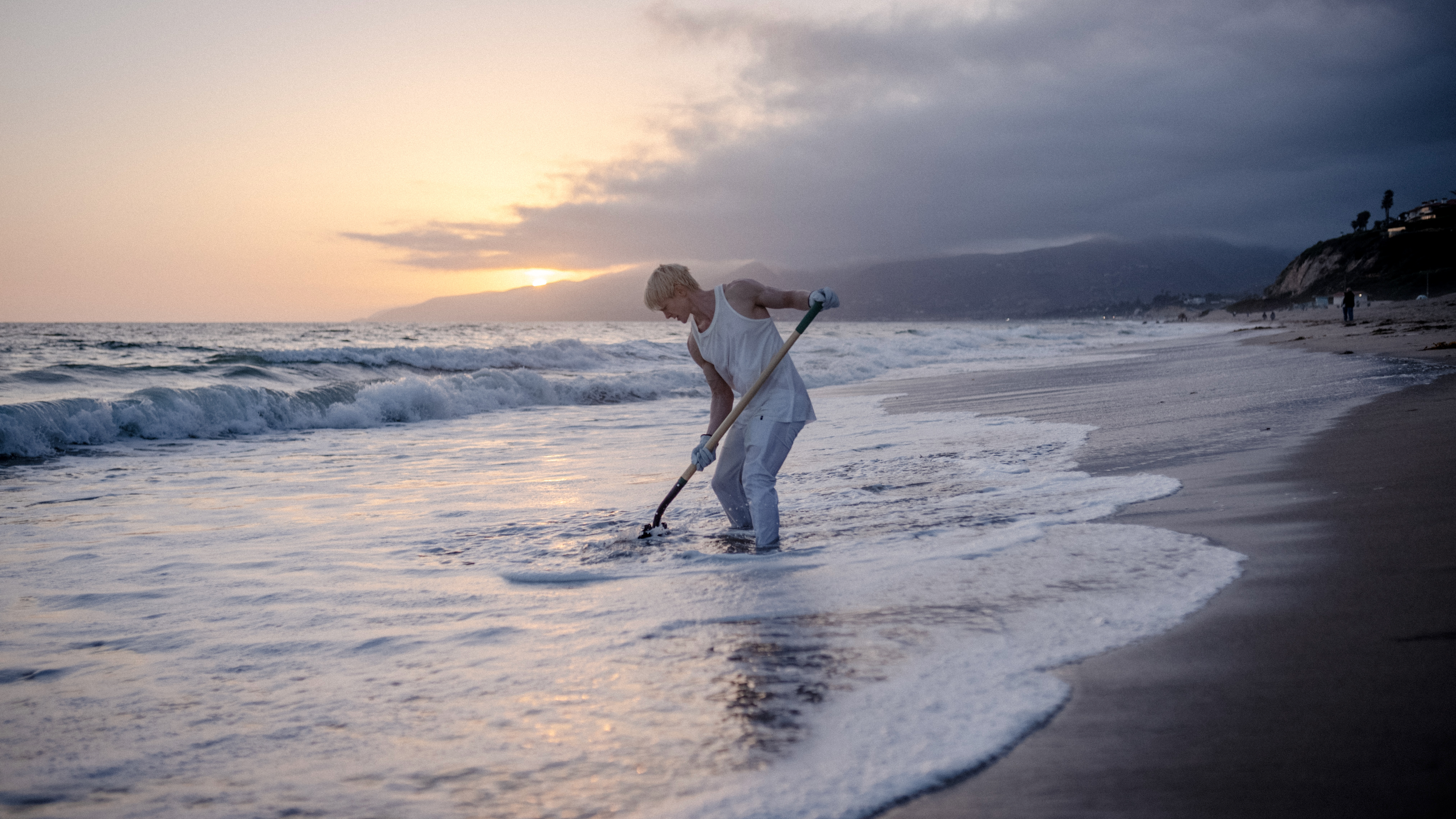 A photograph of a man in a white tank top shovels on the ocean shore at sunrise