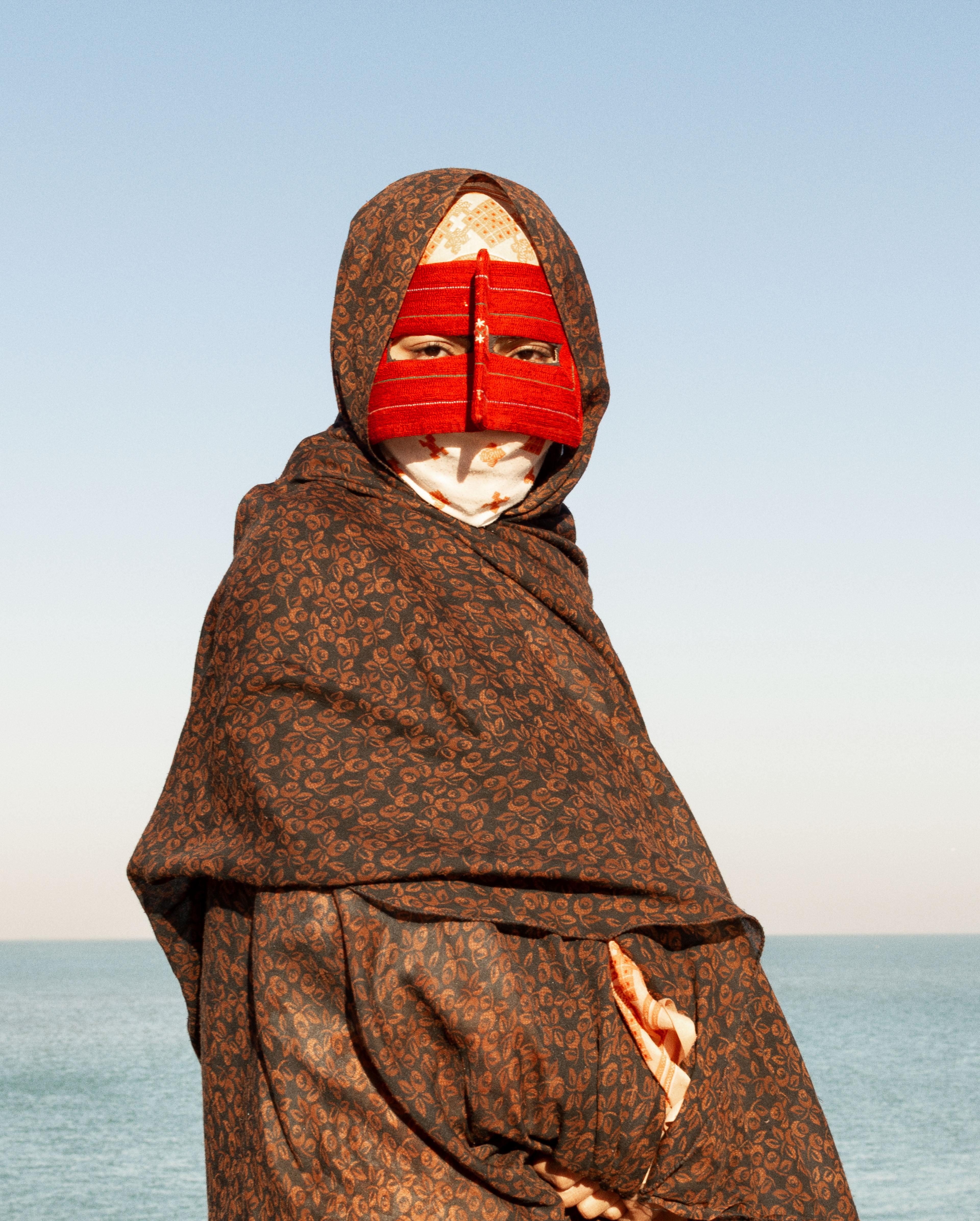 En person står invirad i en sjal och mask över ansiktet, kollar rakt in i kameran