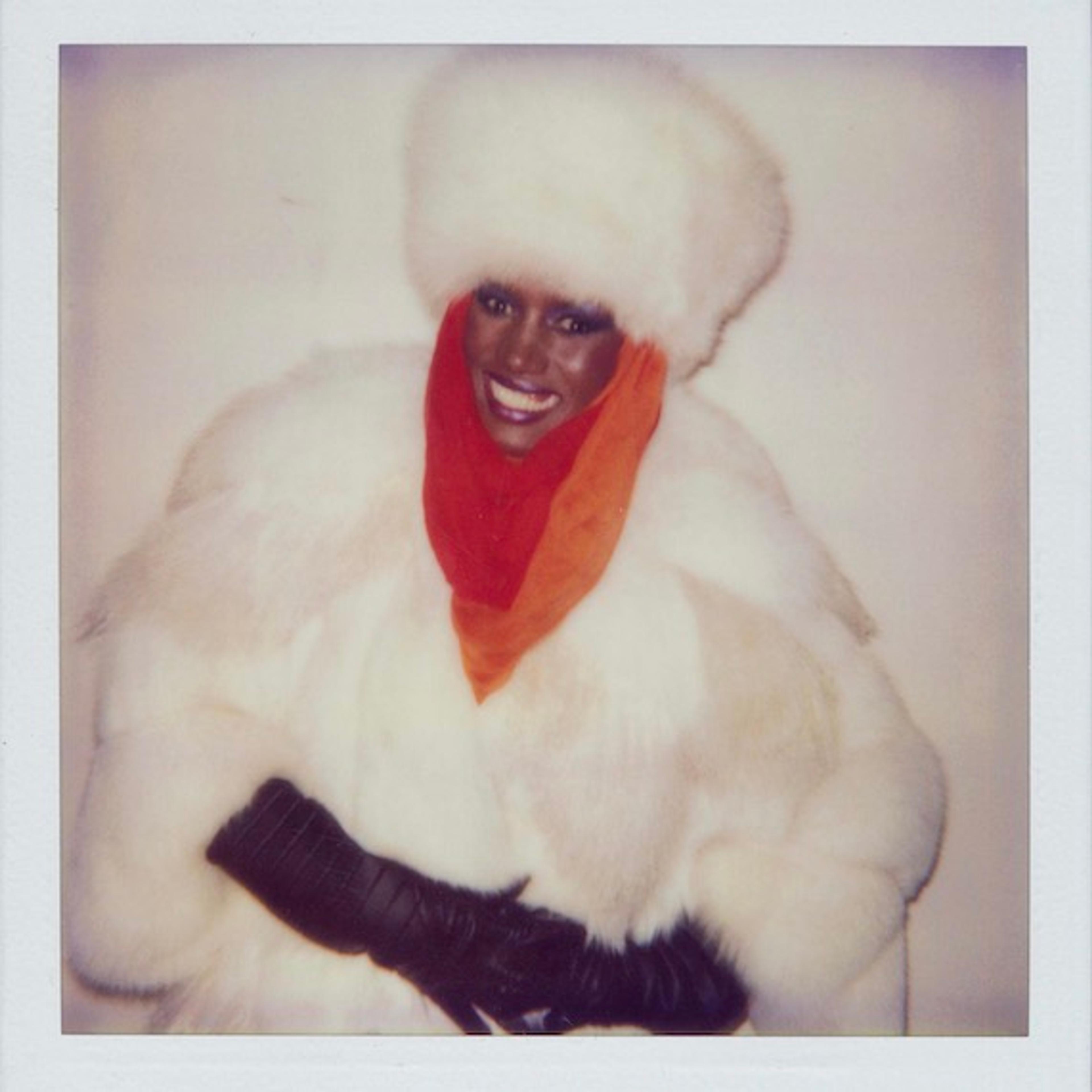A Polaroid photograph of Grace Jones smiling against a plain background, in a white fur coat and hat