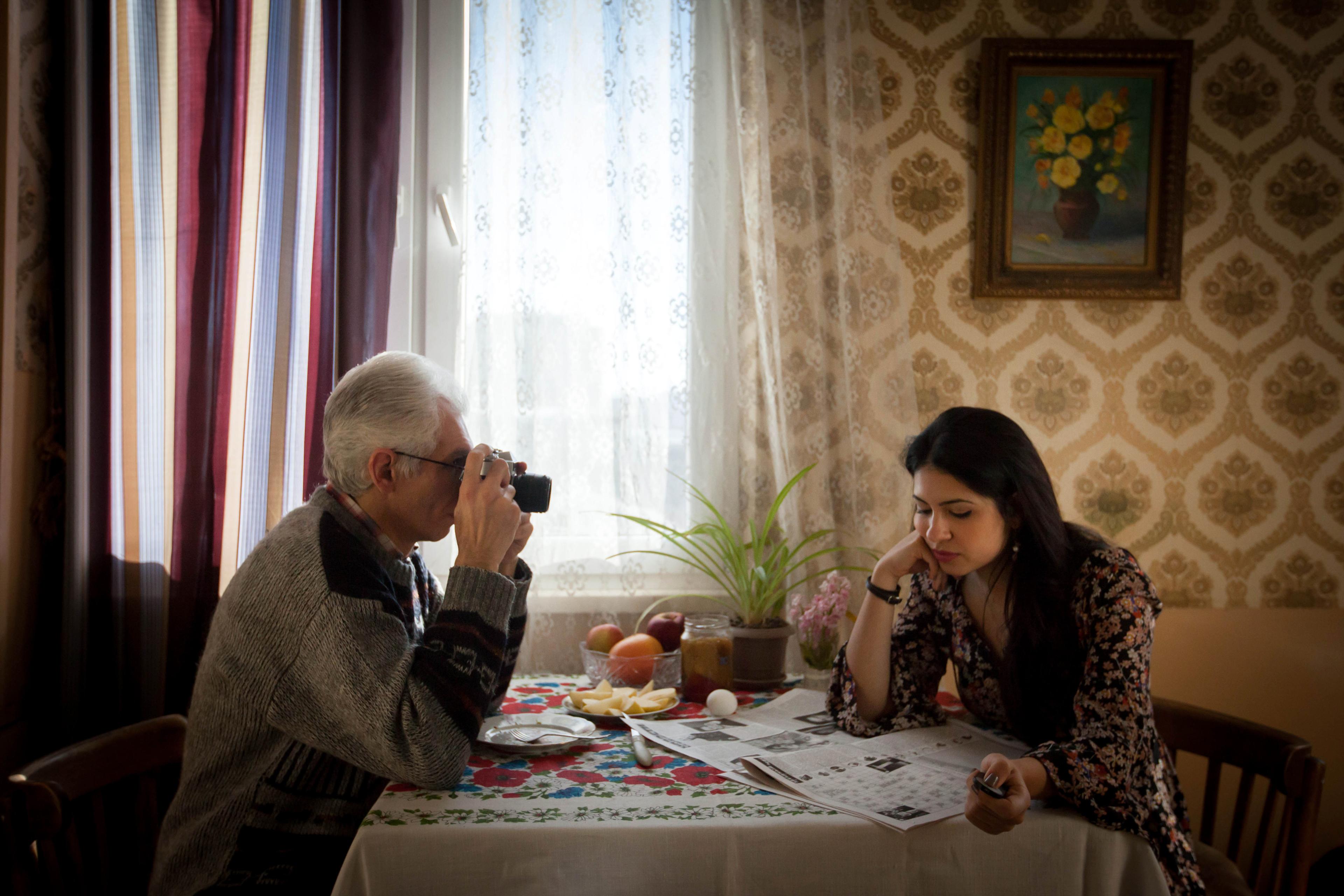 Diana Markosian and her father are sitting at a table. Diana is reading a newspaper, and her father is taking a photo of her with a camera.
