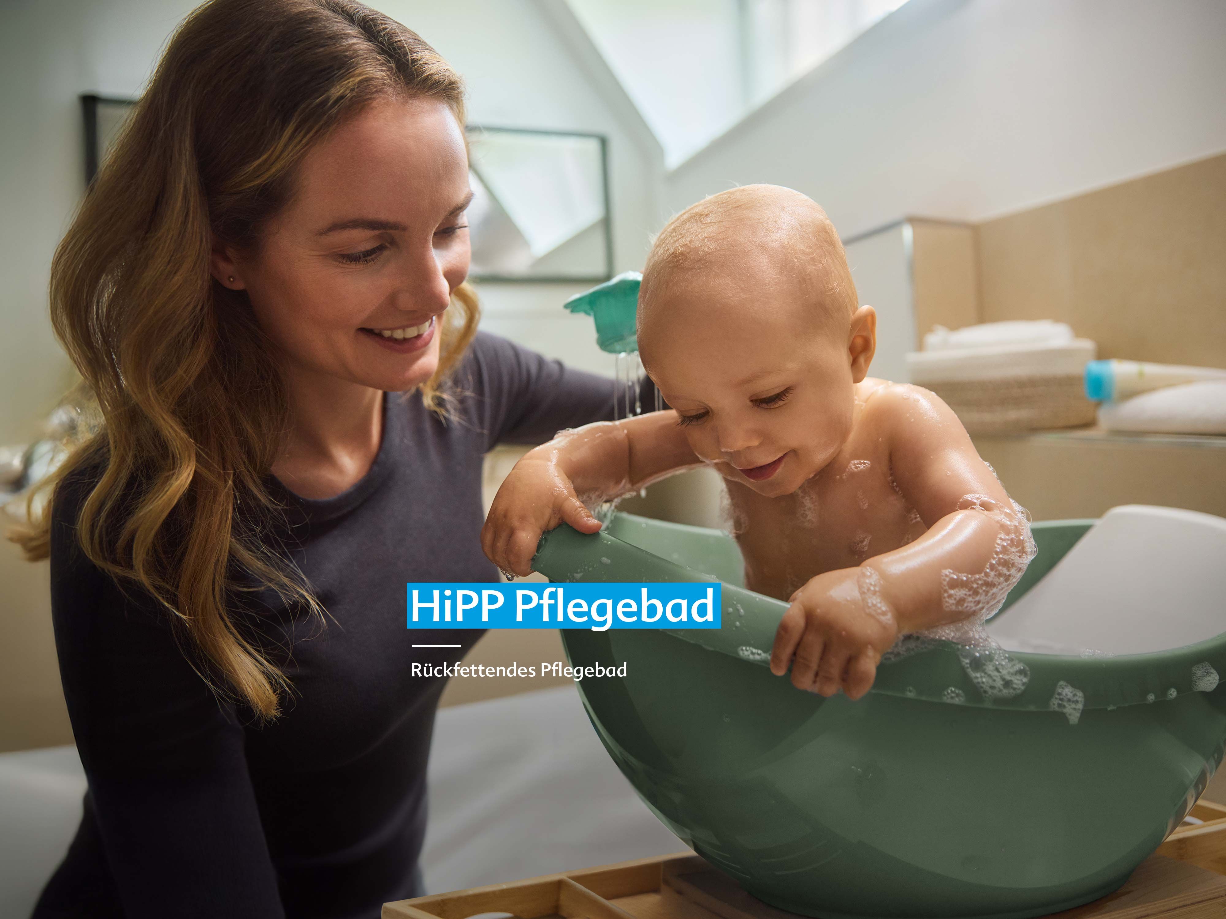 Frau lächelt und hält Baby, das in grünem Babybadewanneneinsatz sitzt und spielt