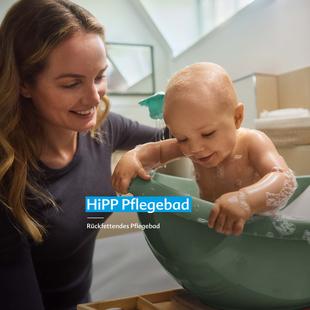 Frau lächelt und hält Baby, das in grünem Babybadewanneneinsatz sitzt und spielt