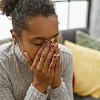 A woman sits on a couch with her face buried in her hands, appearing overwhelmed or upset. She wears a gray sweater and is surrounded by colorful pillows in a cozy living room setting.
