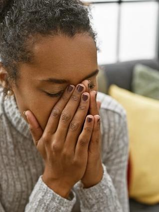A woman sits on a couch with her face buried in her hands, appearing overwhelmed or upset. She wears a gray sweater and is surrounded by colorful pillows in a cozy living room setting.