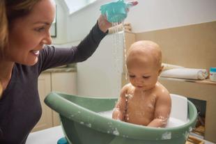 Lachende Mutter badet ihr Baby in einer kleinen Wanne mit Schaum, sanftes Wasser läuft über den Körper im hellen Badezimmer.