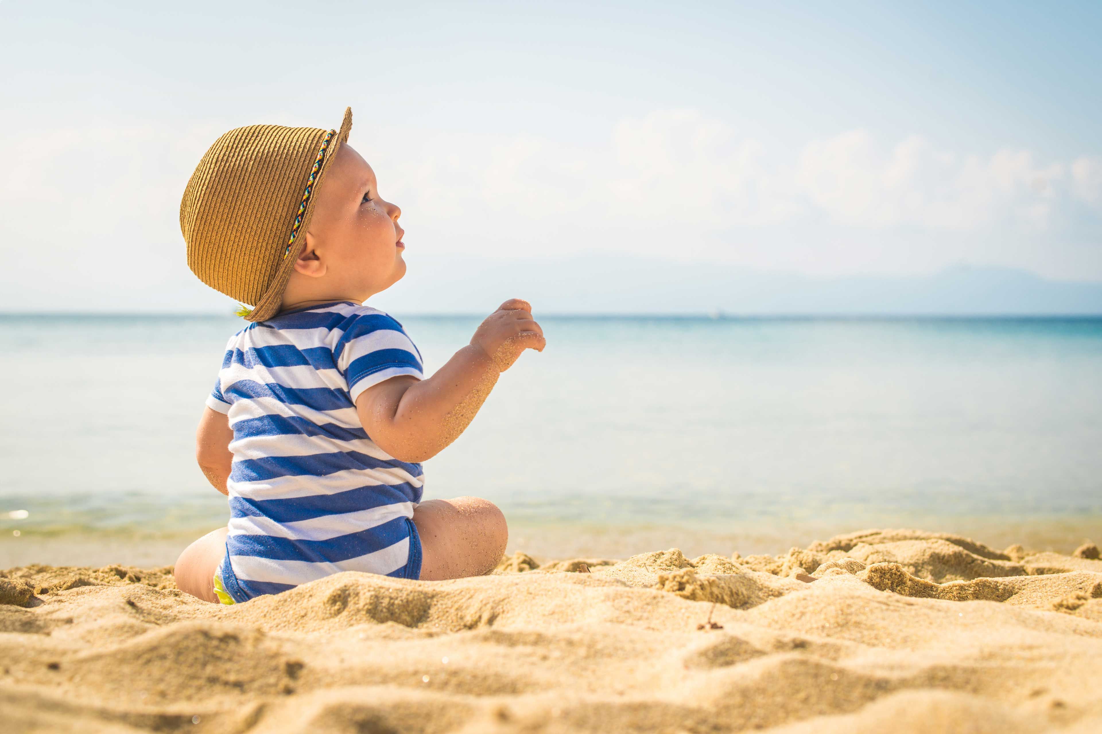 Ein kleines Kind in einem gestreiften Outfit und mit Hut sitzt an einem Sandstrand und blickt mit freudiger Miene in die Ferne.