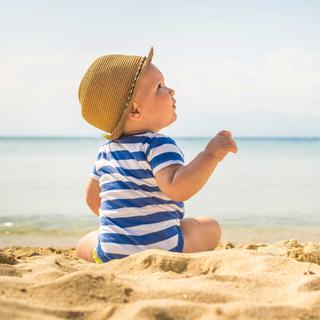 Ein kleines Kind in einem gestreiften Outfit und mit Hut sitzt an einem Sandstrand und blickt mit freudiger Miene in die Ferne.