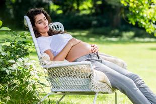 Eine Frau in einer ruhigen Umgebung im Freien, die sanft ihren schwangeren Bauch berührt, während sie auf einem Korbsessel sitzt.