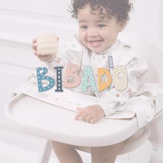 Baby smiling in a highchair wearing a Bibado coverall weaning bib and holding a cup, with the Bibado logo overlaid on the image.