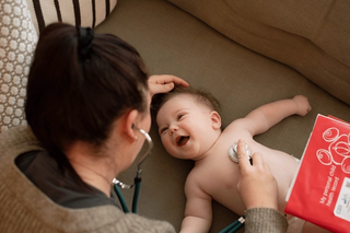 A smiling baby lies on a couch while a woman, likely a healthcare provider, uses a stethoscope to listen to the baby's chest. A red health record booklet labeled "My personal child health record" is visible in the foreground