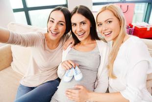 Drei Frauen, von denen zwei Babyschuhe in der Hand halten, lächeln und posieren für ein Selfie.