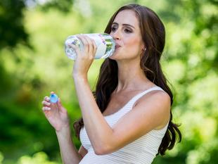 Eine Frau trinkt Wasser aus einer durchsichtigen Flasche, während sie in der anderen Hand einen blau-weißen Gegenstand hält.