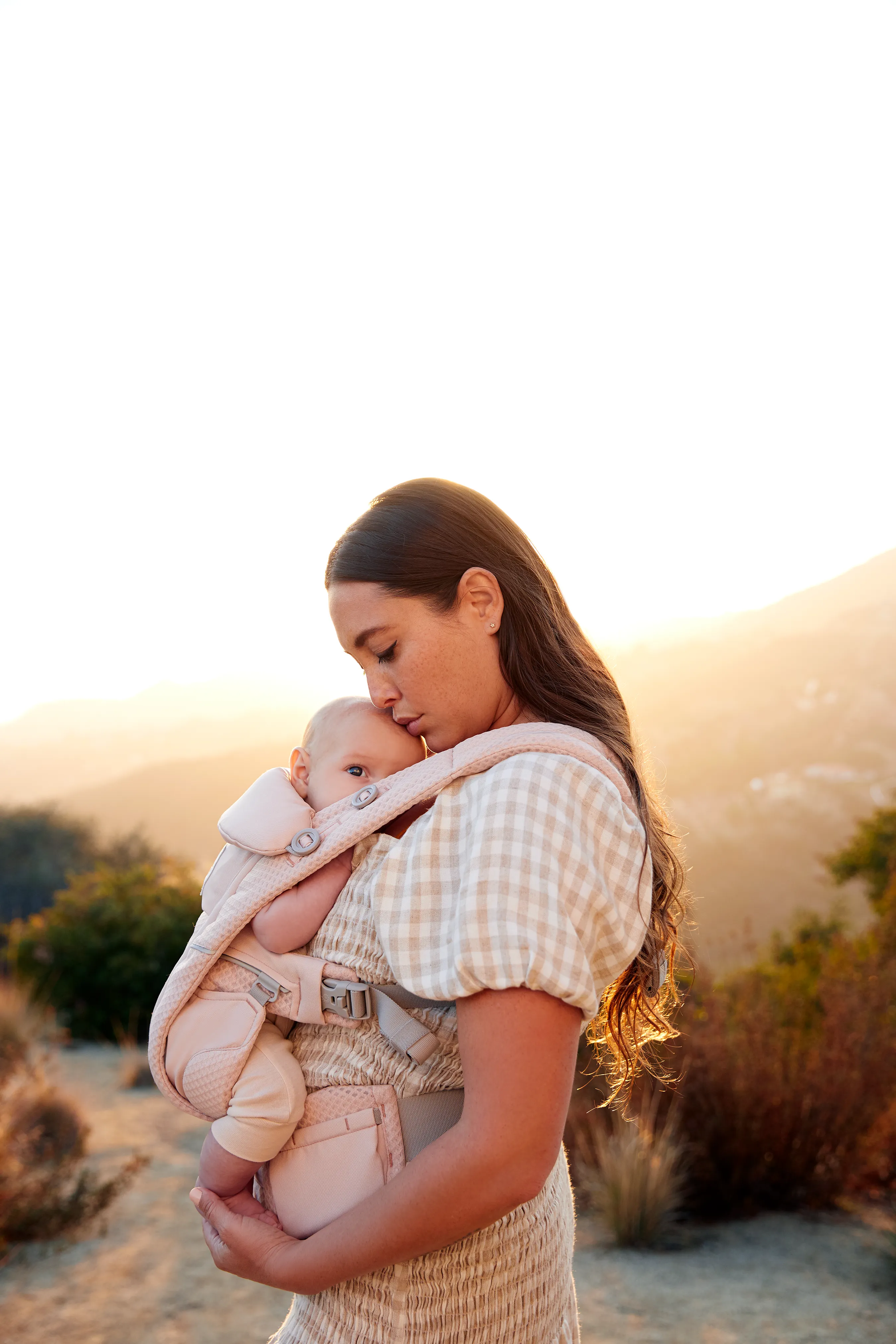 Parent holding baby in a soft pink Ergobaby carrier outdoors at sunset, sharing a quiet, close cuddle.