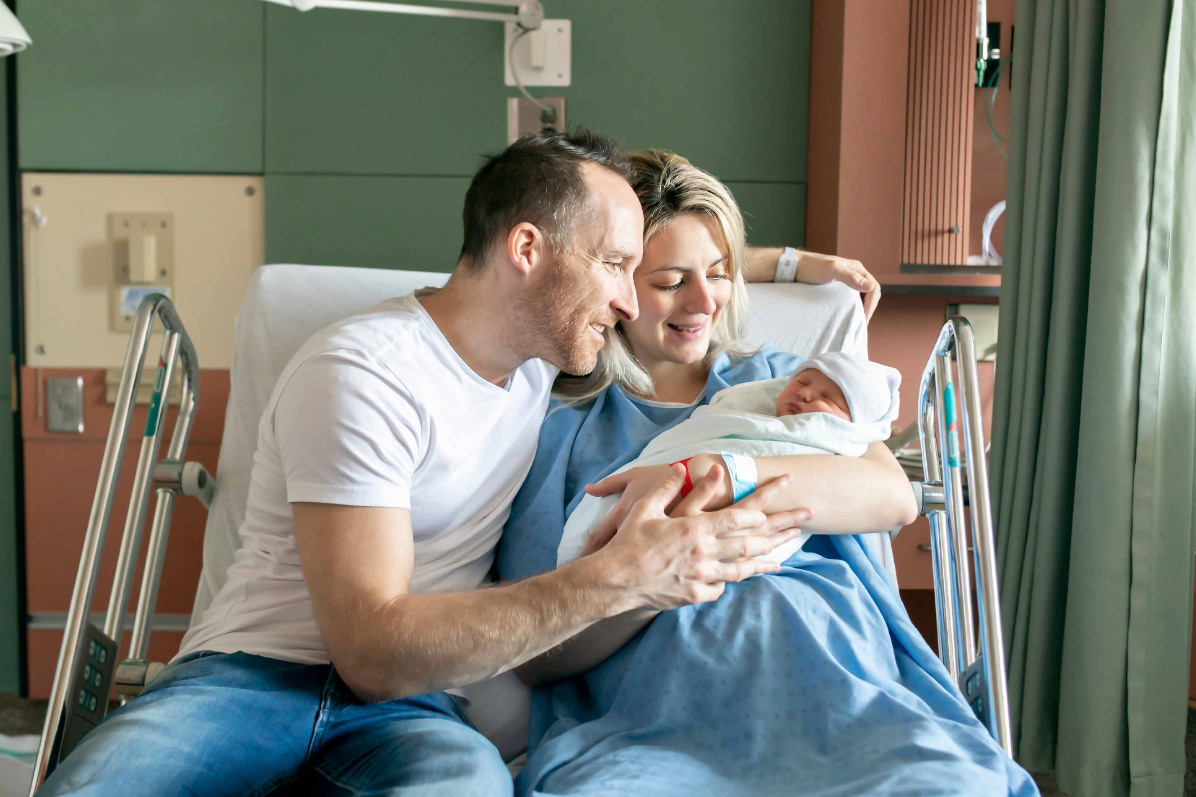 Ein freudiger Moment in einem Krankenhauszimmer, in dem ein Mann und eine Frau ein neugeborenes Baby im Arm halten, beide lächeln und teilen einen intimen Moment.