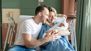 Ein freudiger Moment in einem Krankenhauszimmer, in dem ein Mann und eine Frau ein neugeborenes Baby im Arm halten, beide lächeln und teilen einen intimen Moment.
