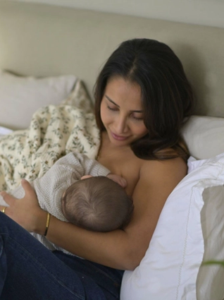 A mother with long dark hair sits on a bed, breastfeeding her baby while wrapped in a soft floral blanket. She gazes down at her child with a gentle smile, her arm securely cradling the infant. The cozy setting includes plush white pillows and a neutral-toned headboard, creating a warm and serene atmosphere.