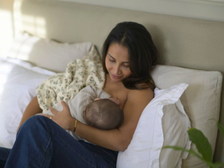 A mother with long dark hair sits on a bed, breastfeeding her baby while wrapped in a soft floral blanket. She gazes down at her child with a gentle smile, her arm securely cradling the infant. The cozy setting includes plush white pillows and a neutral-toned headboard, creating a warm and serene atmosphere.
