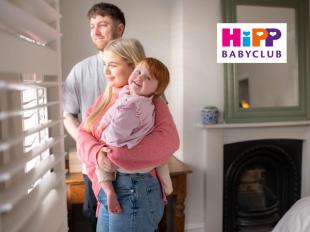 Promotional image for HiPP Babyclub featuring a young family—mother, father, and their red-haired toddler—smiling together in a bright, modern home.
