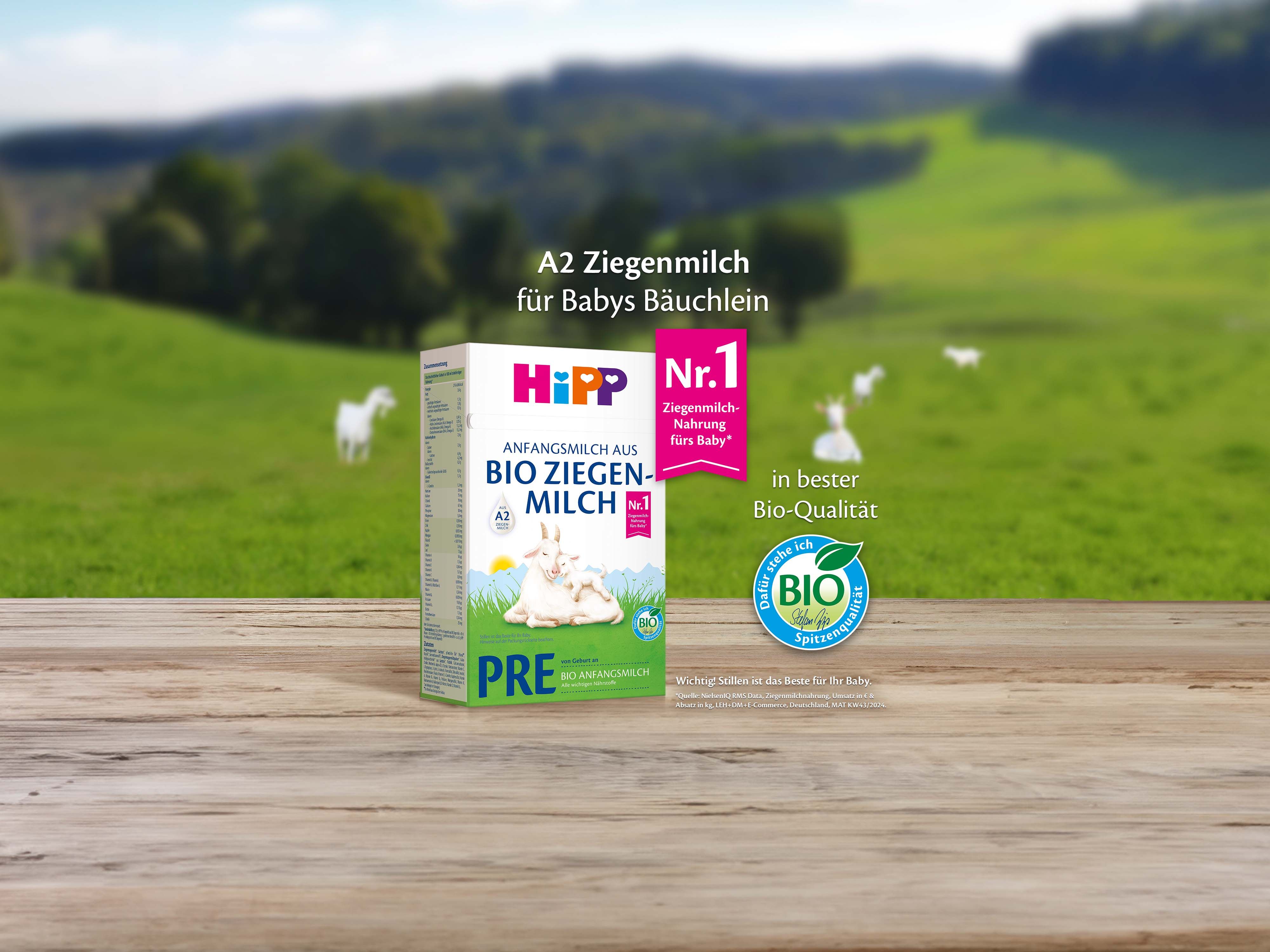 Packung HiPP Bio Ziegenmilch PRE mit Bio-Siegel auf Holztisch vor grüner Landschaft mit Ziegen im Hintergrund