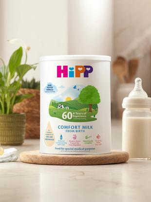 HiPP Comfort milk tub on a bright kitchen worktop beside a prepared baby bottle, dummy, muslin cloth, plant and soft toy
