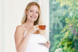 Eine Frau mit blondem Haar, die eine Tasse Tee in der Hand hält, steht an einem Fenster mit Blick ins Grüne.