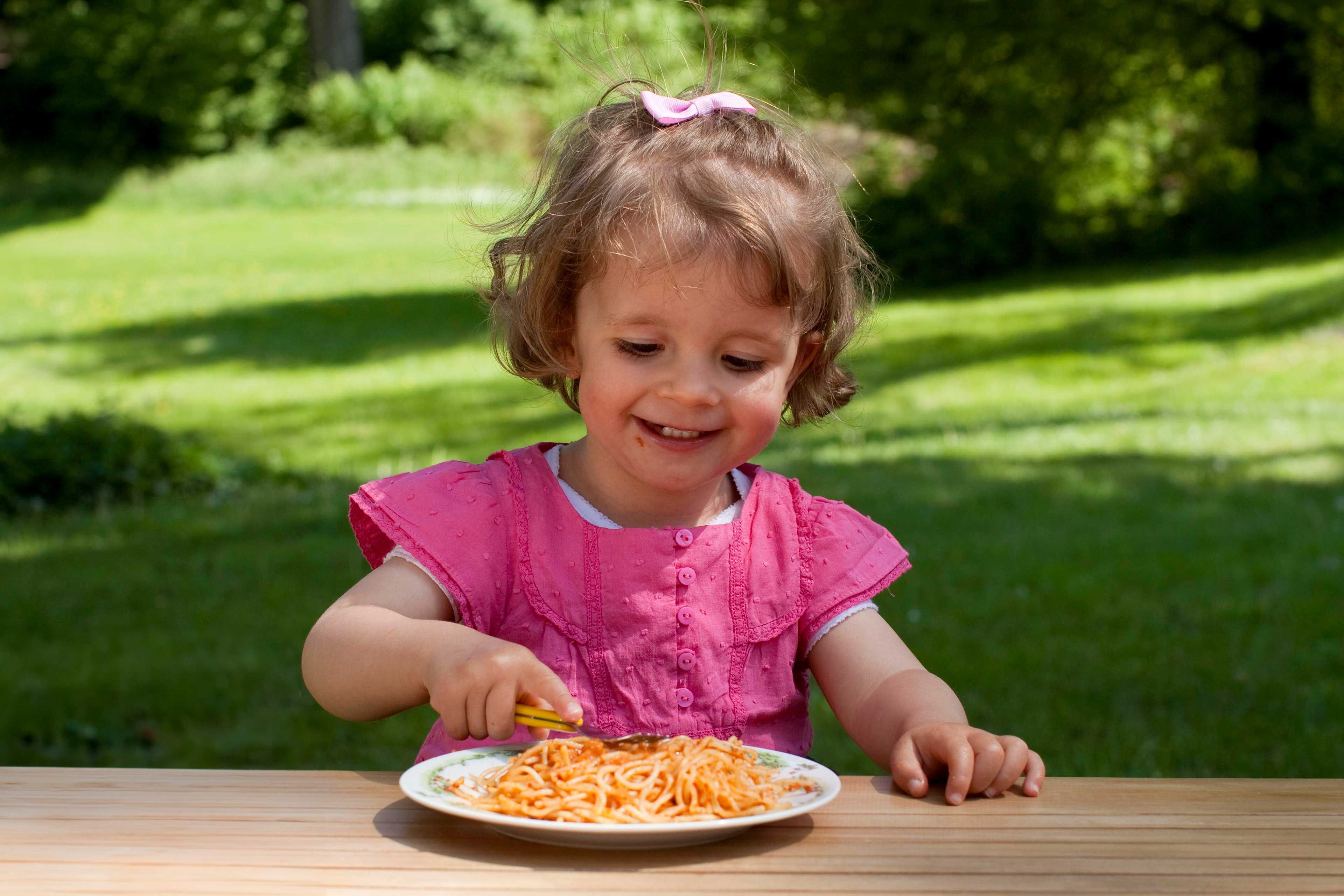 Kleinkind in rosa Kleid sitzt an Holztisch im Freien und isst Spaghetti von einem Teller