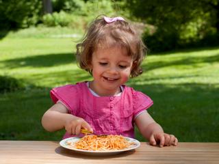 Kleinkind in rosa Kleid sitzt an Holztisch im Freien und isst Spaghetti von einem Teller